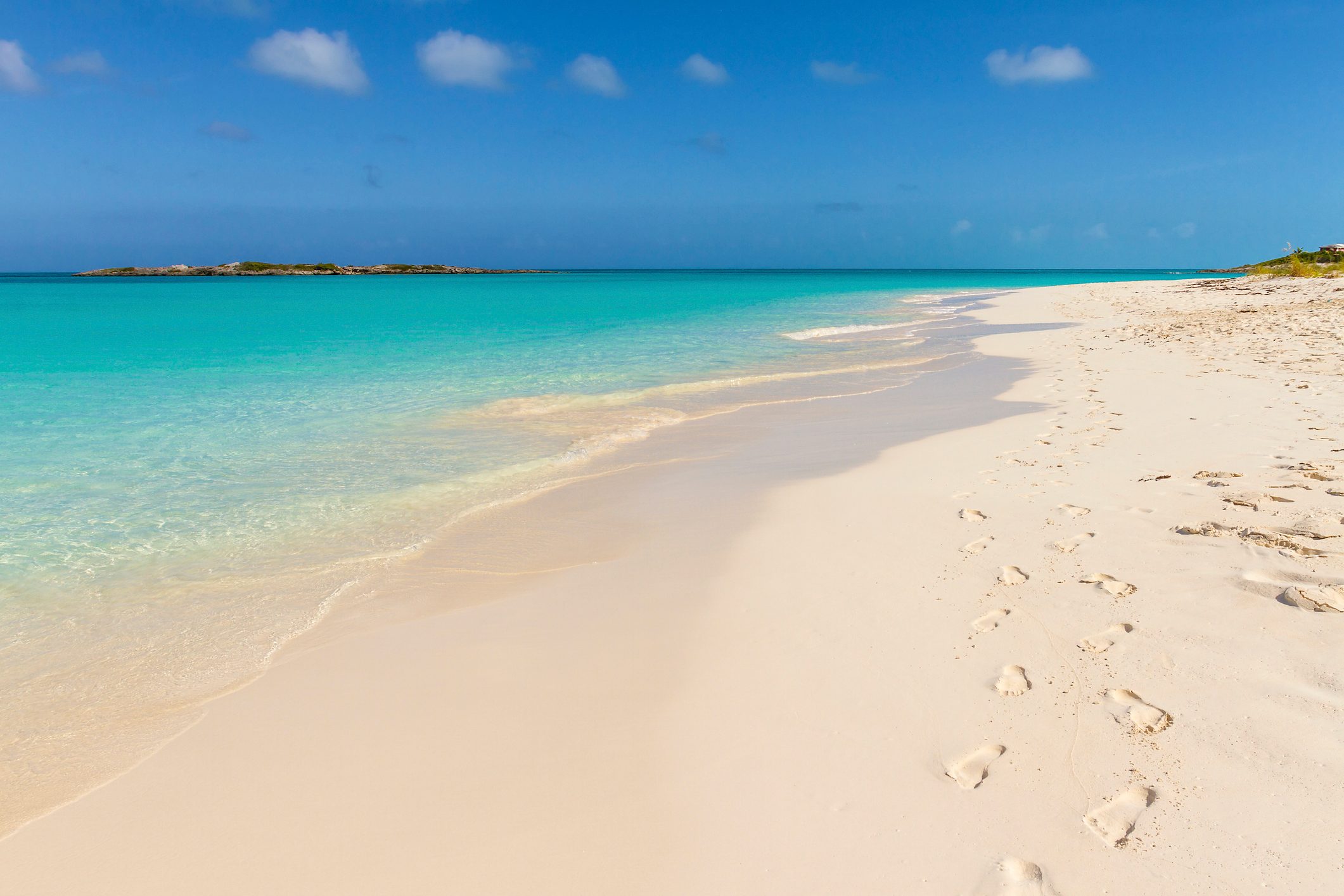 Footprints trail across a pristine sandy beach beside turquoise water, under a clear blue sky with a distant island horizon.