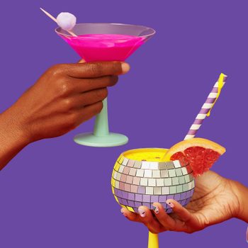 Hands holding brightly colored cocktails; one in a martini glass and the other in a mosaic cup, against purple background.