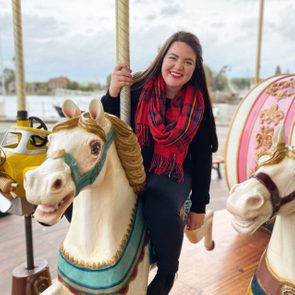 A person rides a carousel horse, smiling, wearing a red plaid scarf, surrounded by other carousel figures, outdoors near a waterfront area.