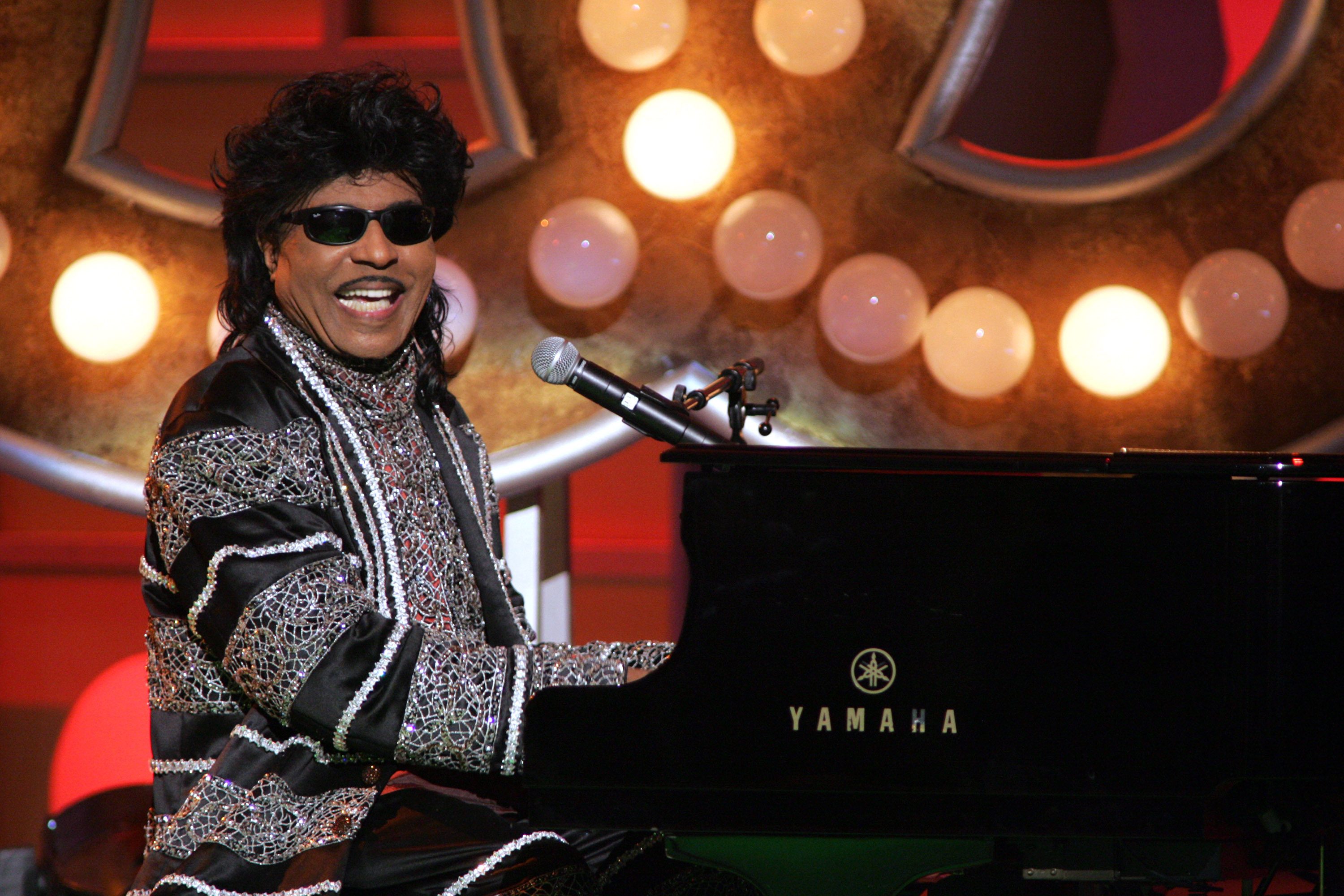 Performer plays Yamaha piano, smiling and wearing intricate sequined attire, in front of illuminated backdrop with circular lights.