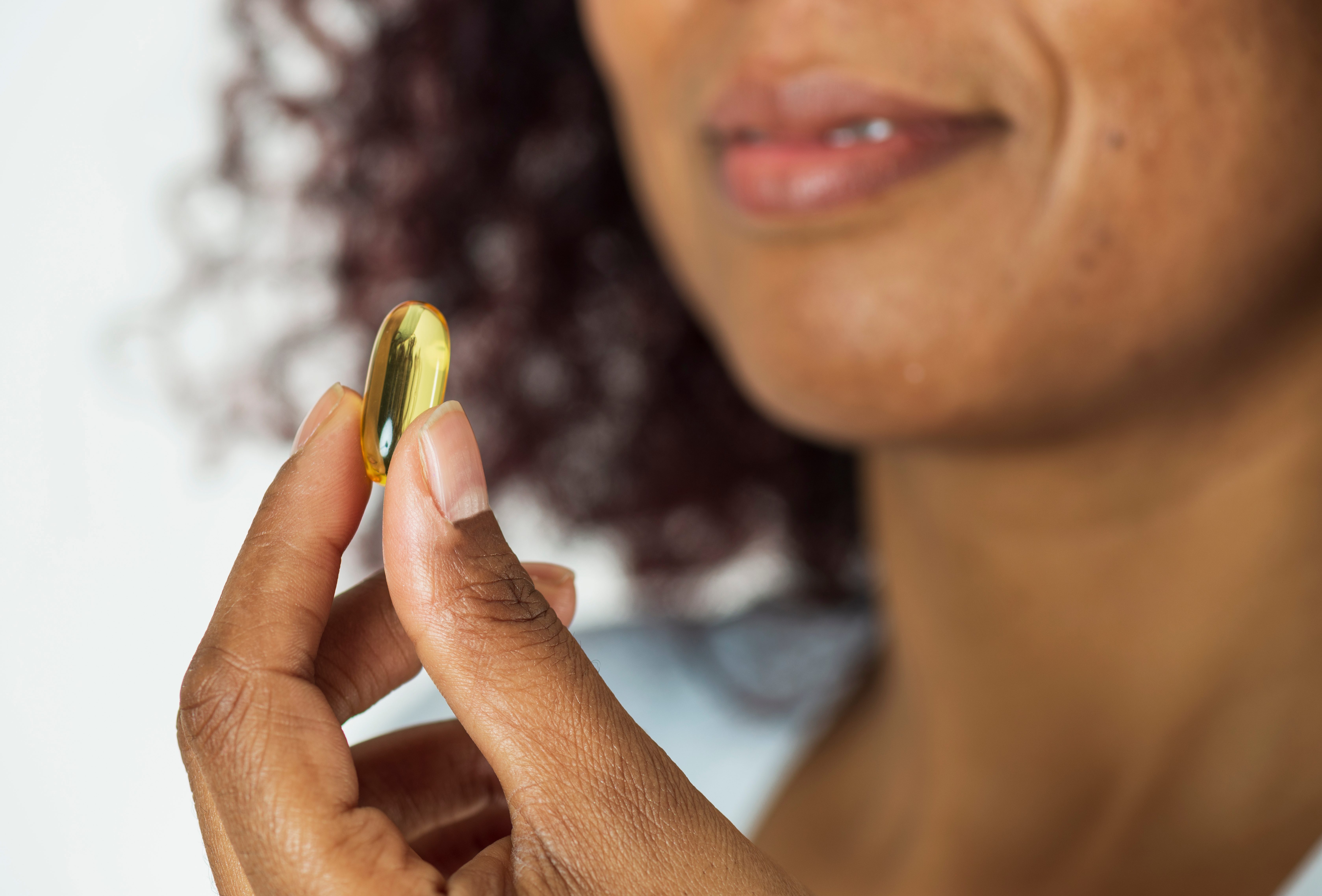 A hand holds a yellow capsule near a woman's face, against a soft-focus background.