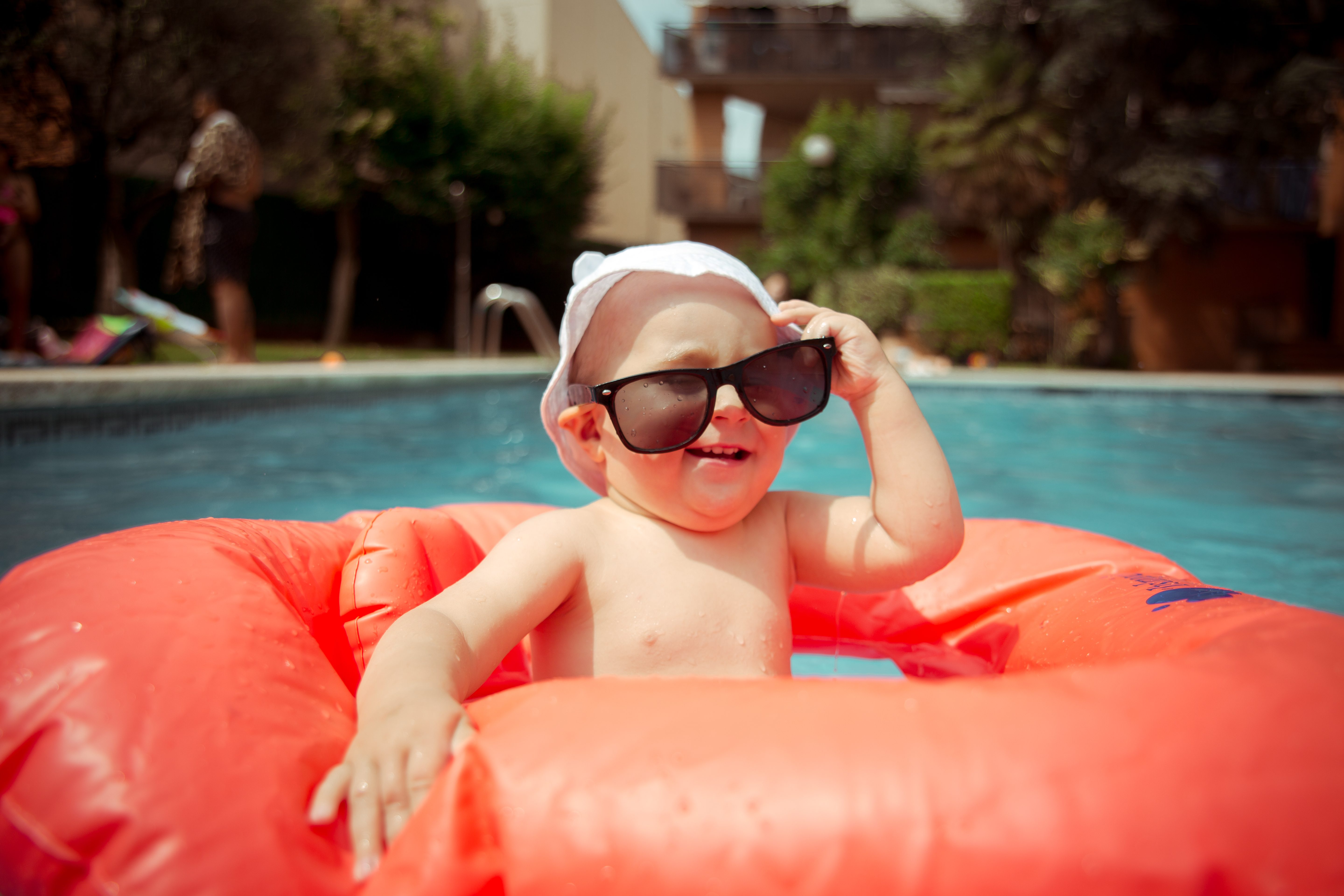 Baby wearing sunglasses smiles while sitting in an orange inflatable float in a sunny poolside environment.