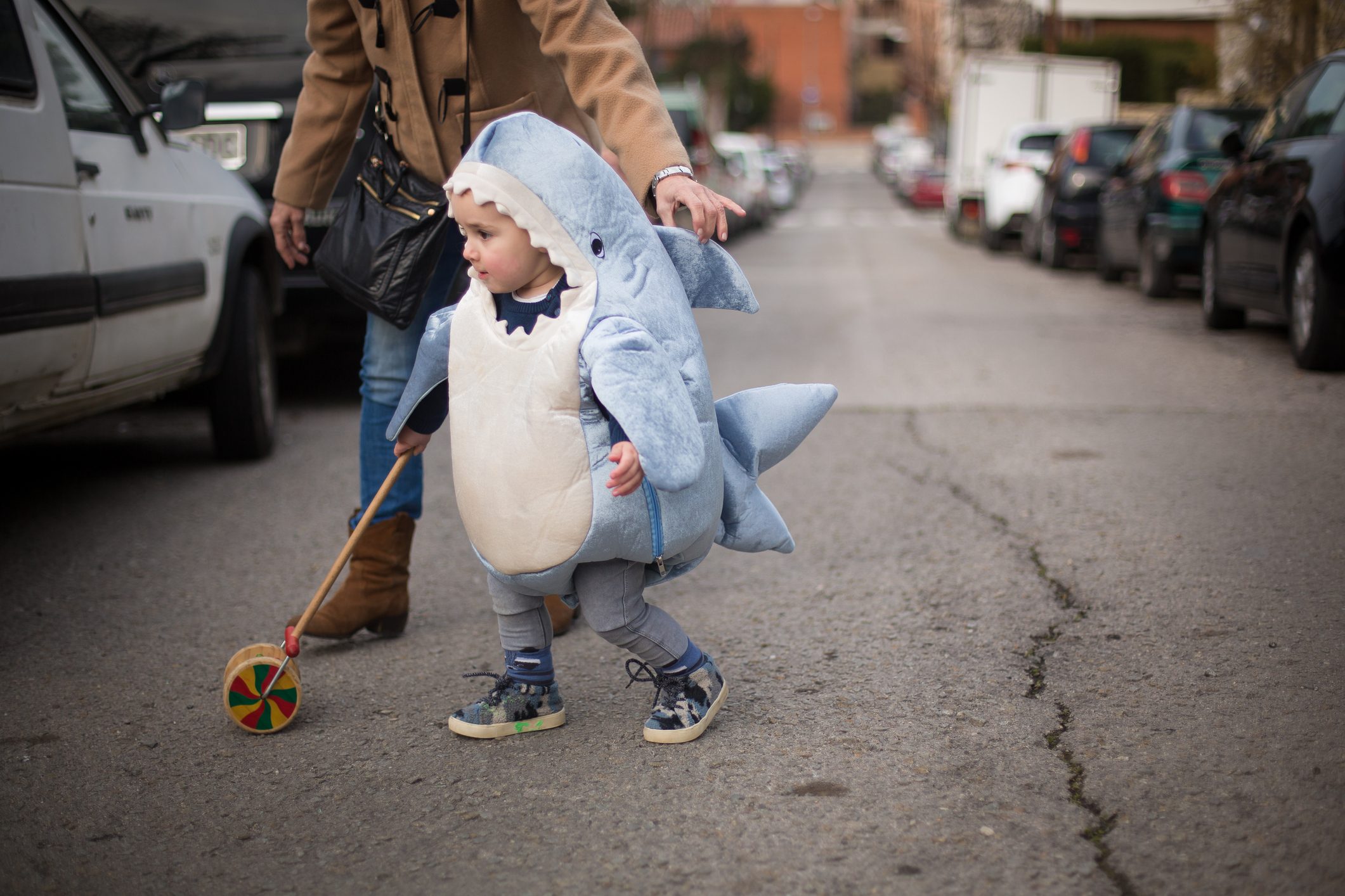 Child in a shark costume walks with a wheeled toy on a street. Nearby adult gently guides the child. Parked cars line the road.