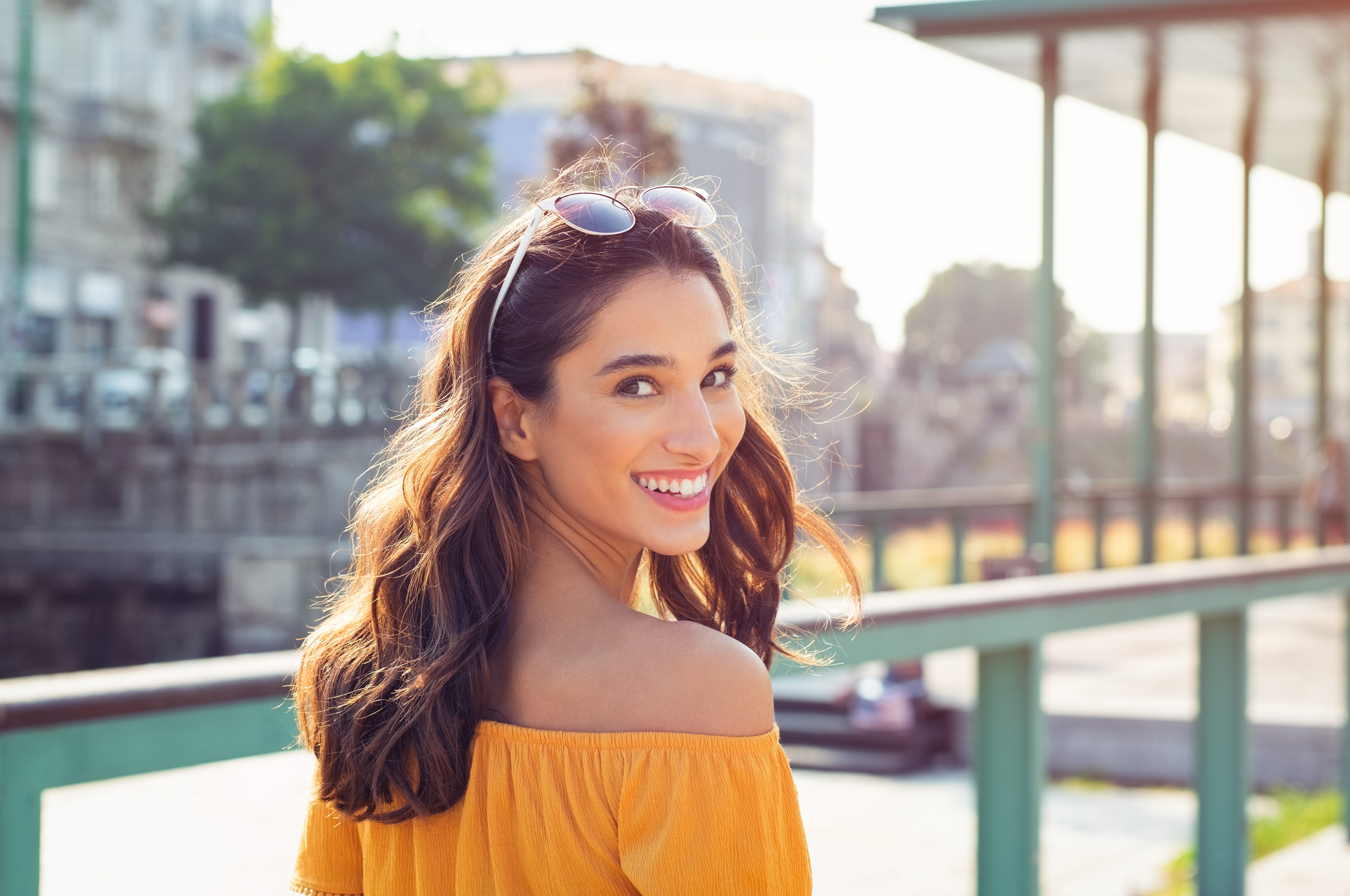 A woman smiles while looking back, wearing sunglasses on her head, in a sunny outdoor urban setting.