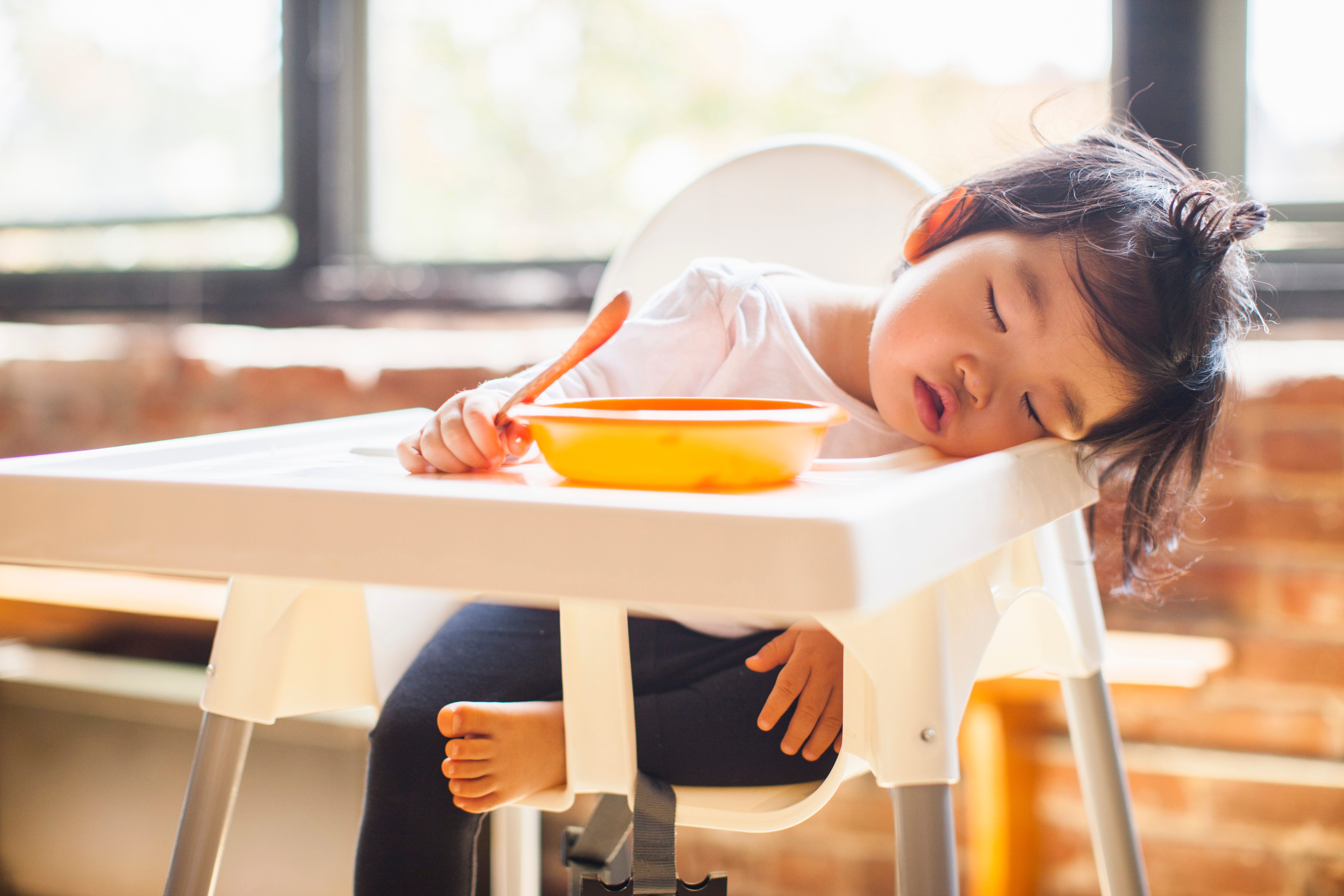 Child sleeps at highchair, head resting beside an orange bowl, in a sunlit room with large windows and bricks.