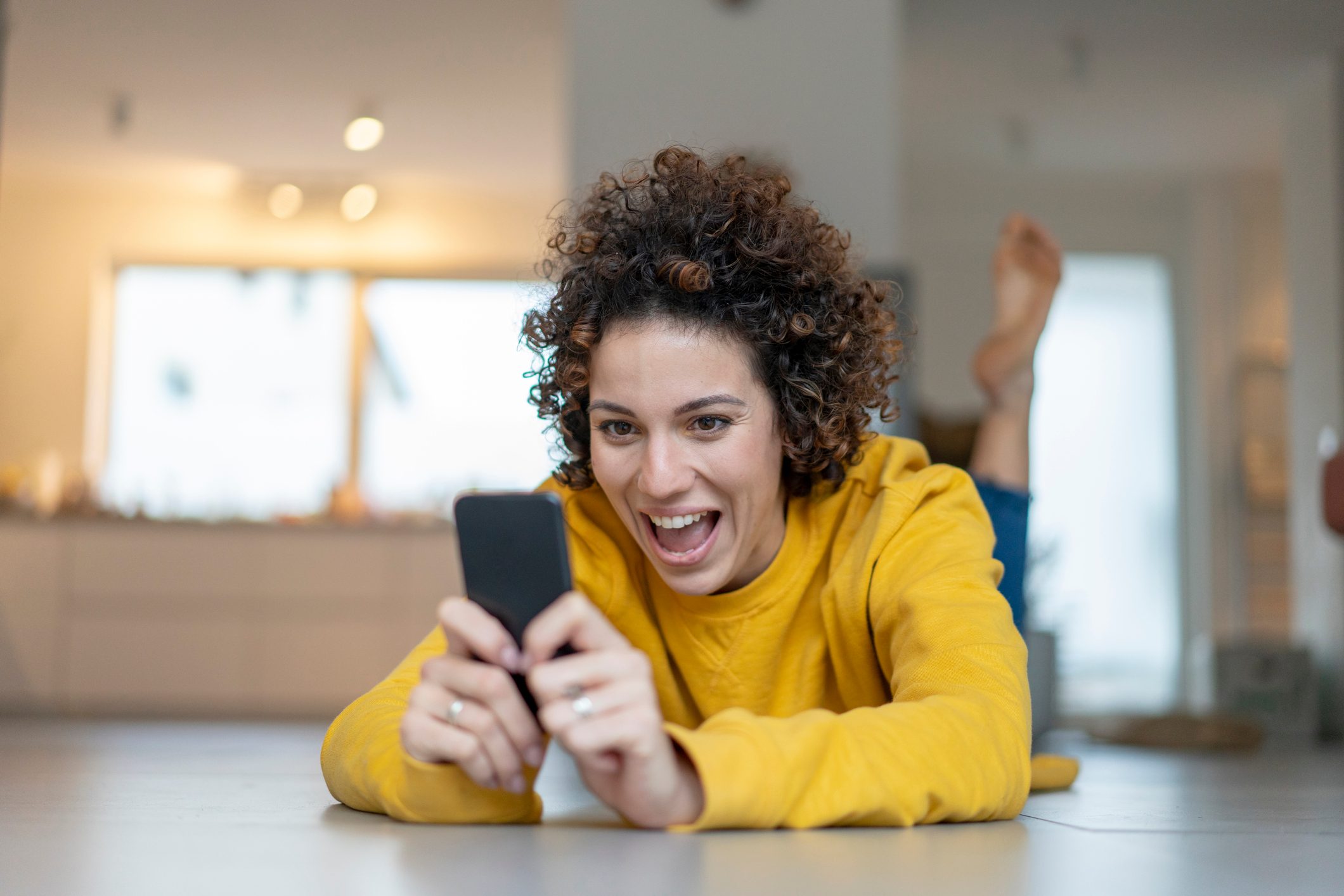 Person smiles, holding a smartphone, lying on the floor in a bright, modern room with soft lighting.