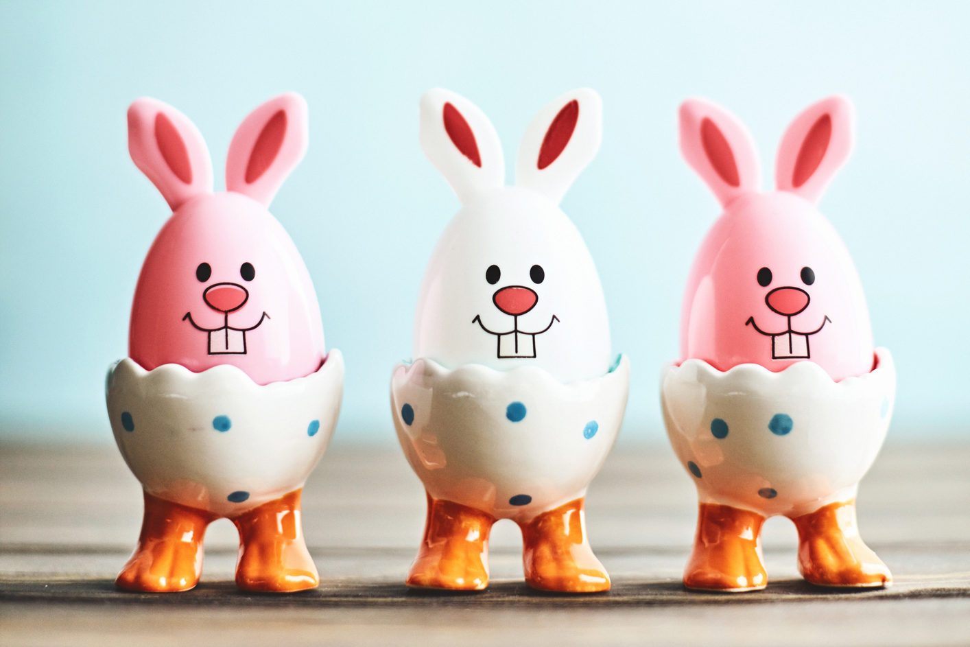 Egg-shaped bunnies rest in dotted egg cups on wooden surface, blue background.