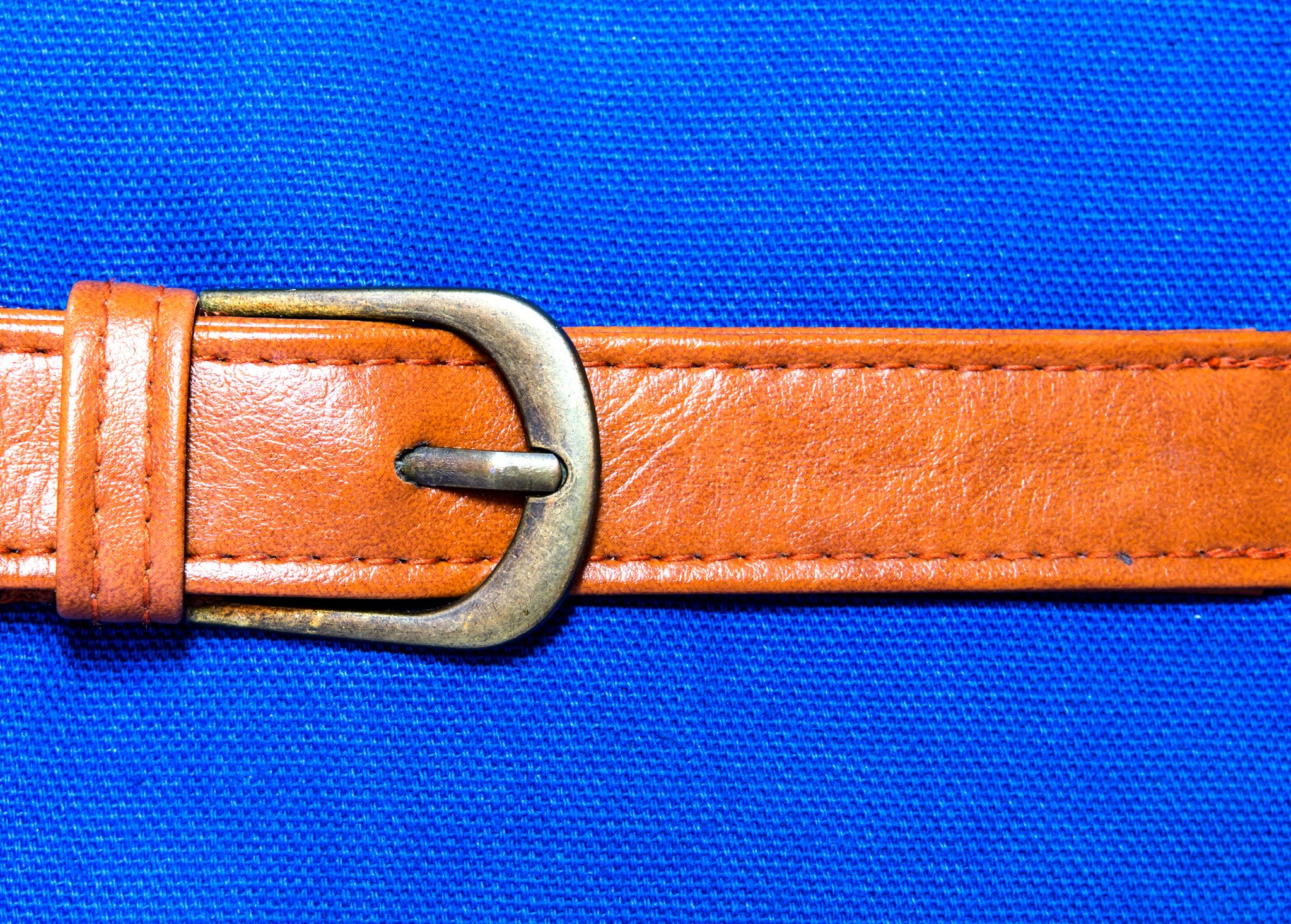 A brown leather belt lies flat with a metal buckle, set against a textured blue fabric background.