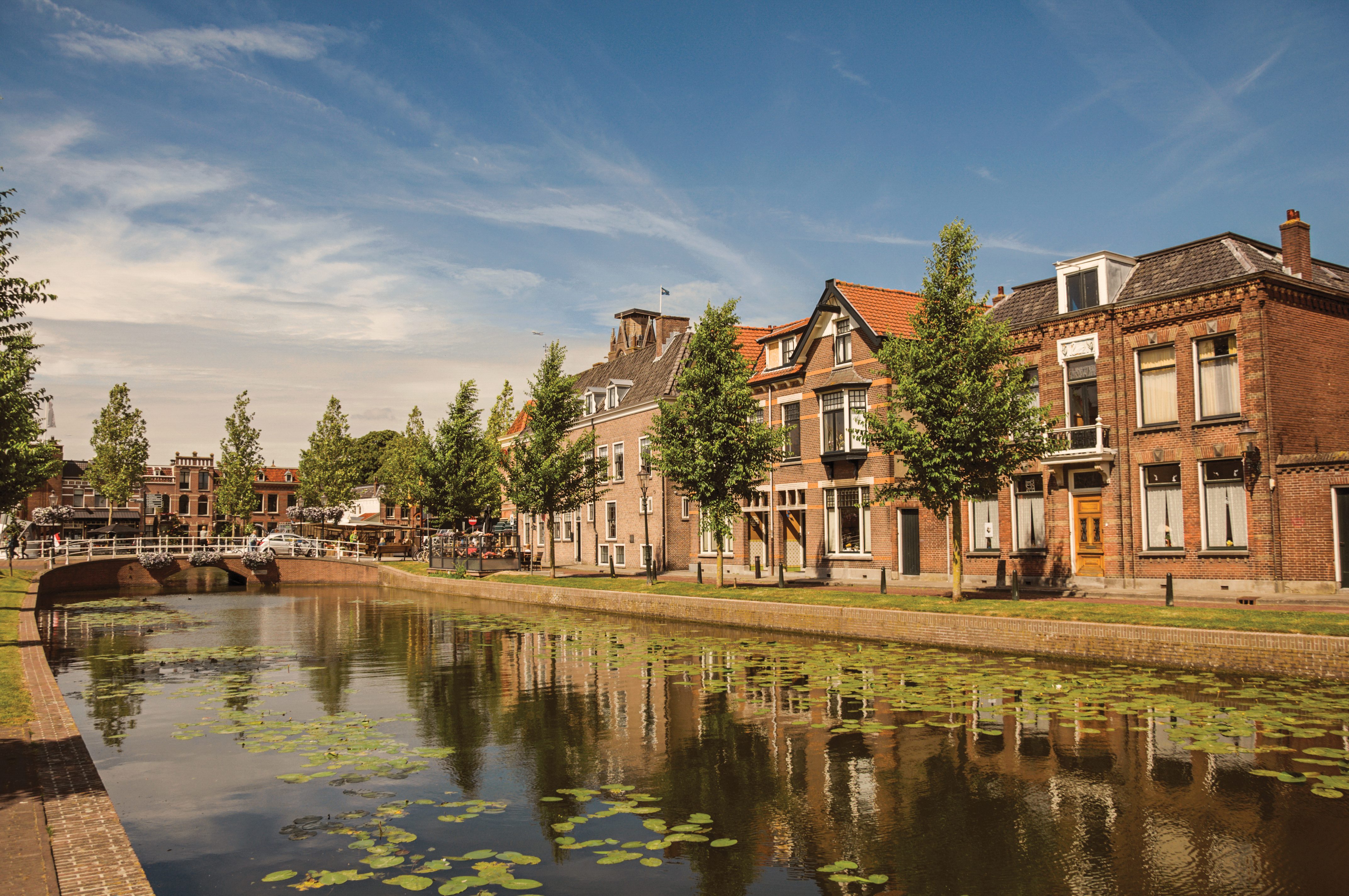 Canal reflects brick buildings and trees, with a bridge in the distance under a clear blue sky.