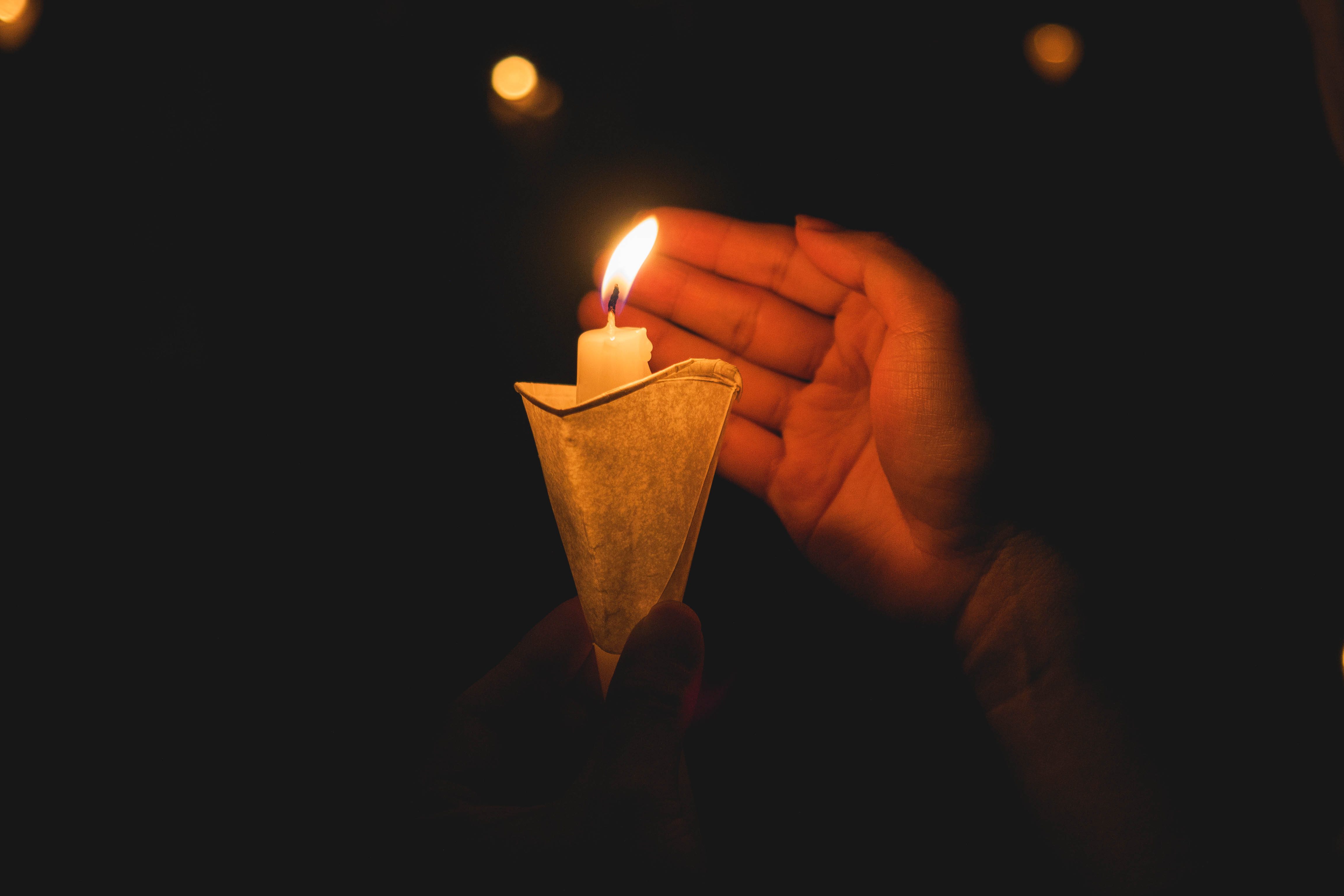 Candle burns gently, shielded by a hand, amid surrounding darkness.