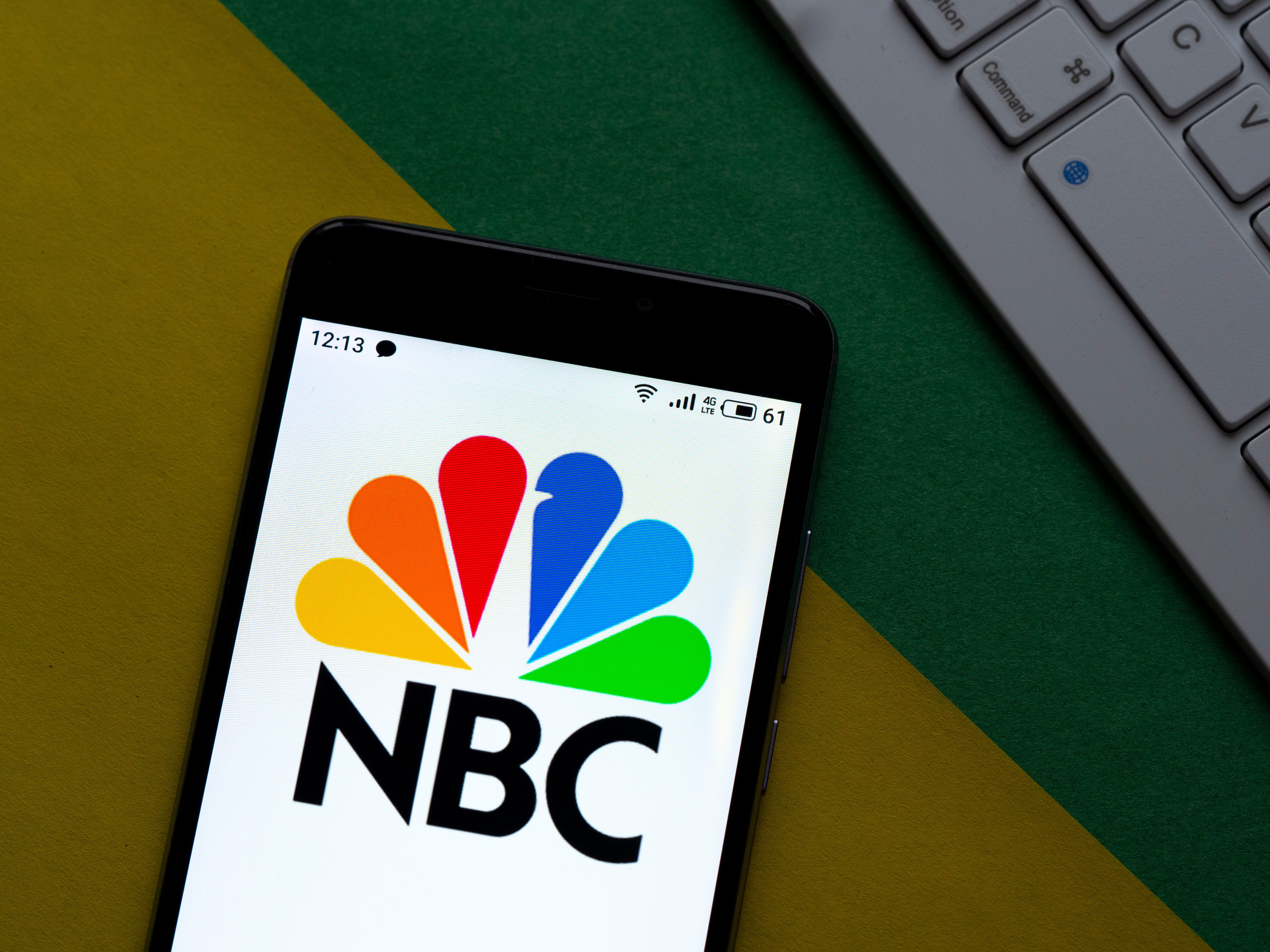 Phone displays NBC logo, colorful peacock feathers, placed on a green and yellow background, next to a keyboard.