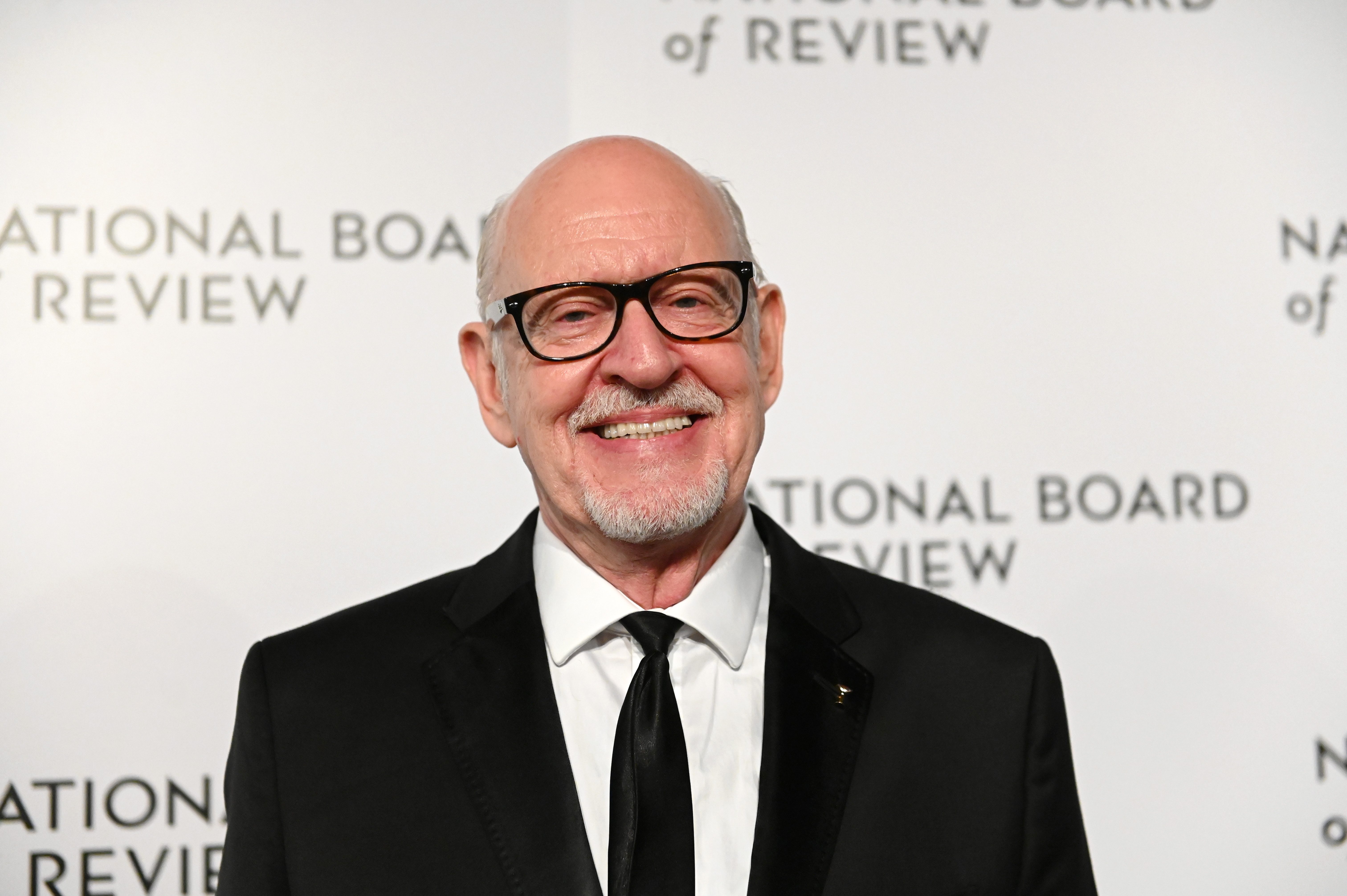 Man smiling, wearing a suit, in front of a "National Board of Review" backdrop at an event.