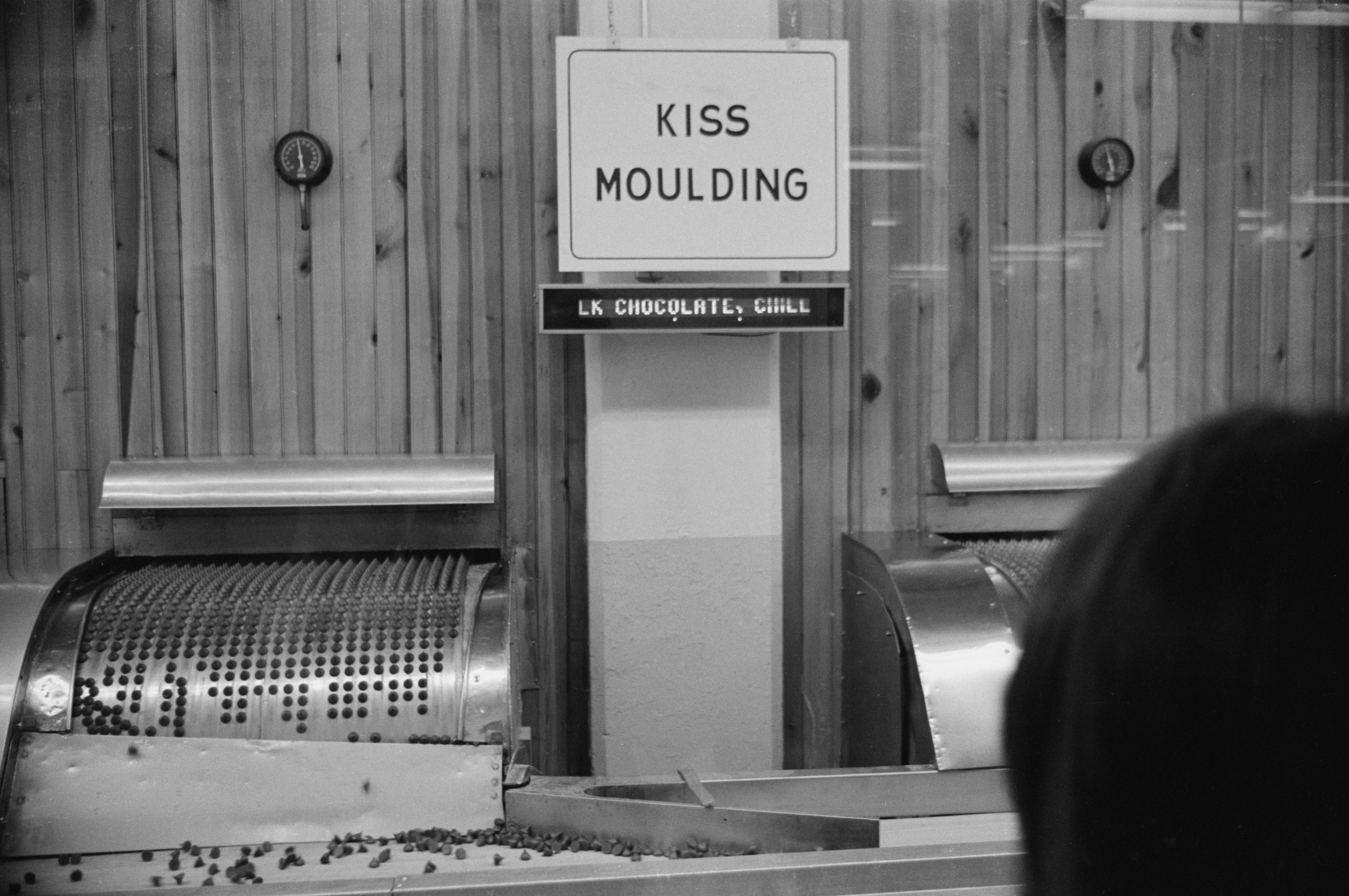 Moulding machine forms chocolates beneath "KISS MOULDING" sign in a wooden-paneled room.