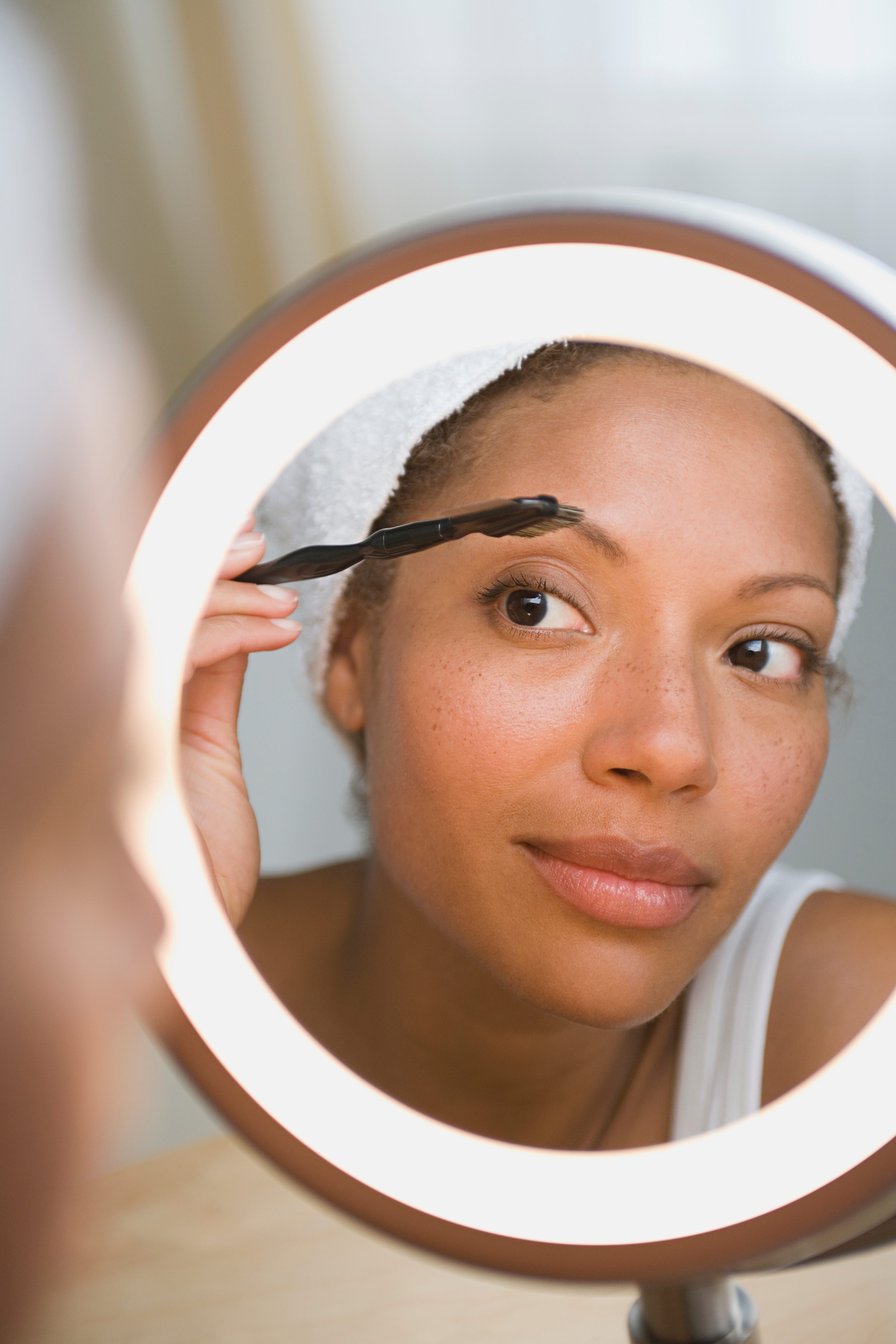 Woman tidies eyebrows using brush, reflected in illuminated circular mirror, indoors.