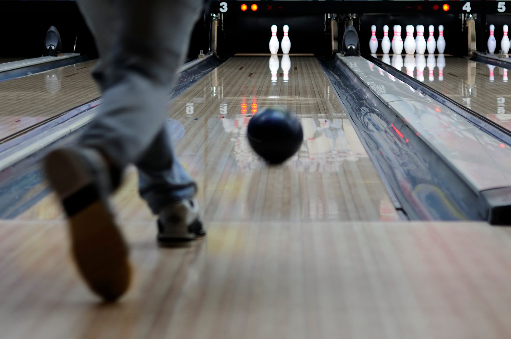 Bowling ball rolls down a lane towards pins, while a person's foot crosses the foul line in a bowling alley.