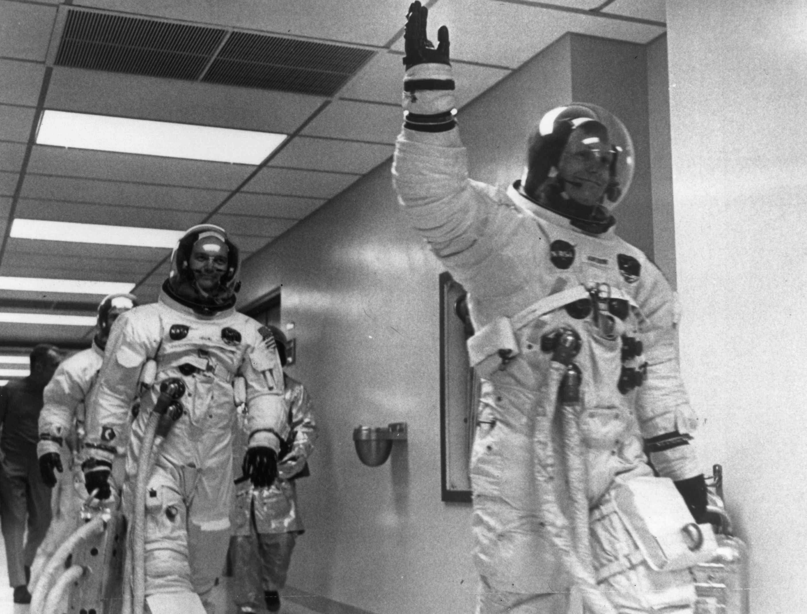 Astronauts walk through hallway, wearing spacesuits, with one raising a hand in greeting. The corridor is brightly lit with overhead panels.