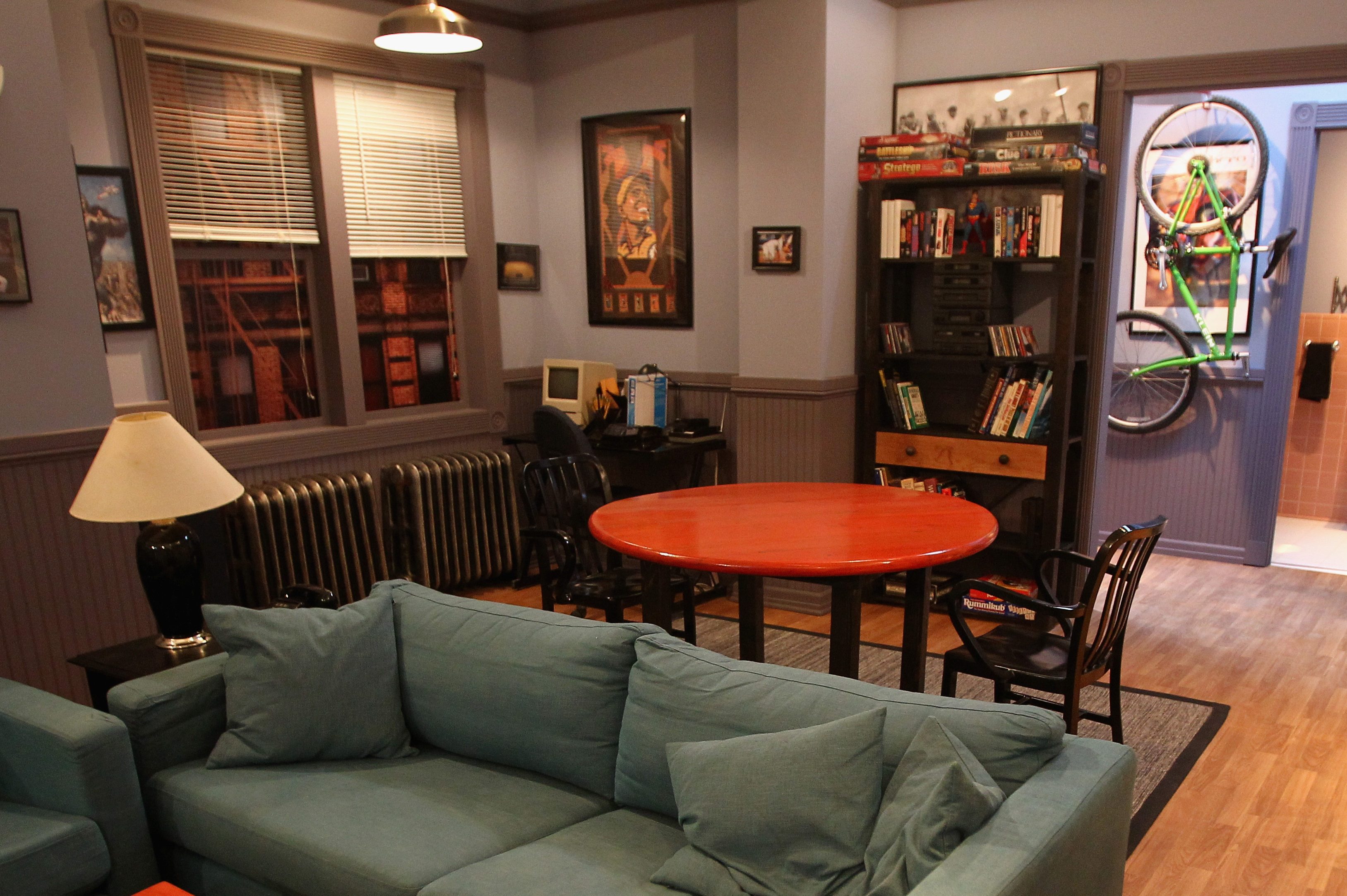 A cozy living room with green sofas, a red round table, bookshelves, a bike on the wall, and a window with blinds.