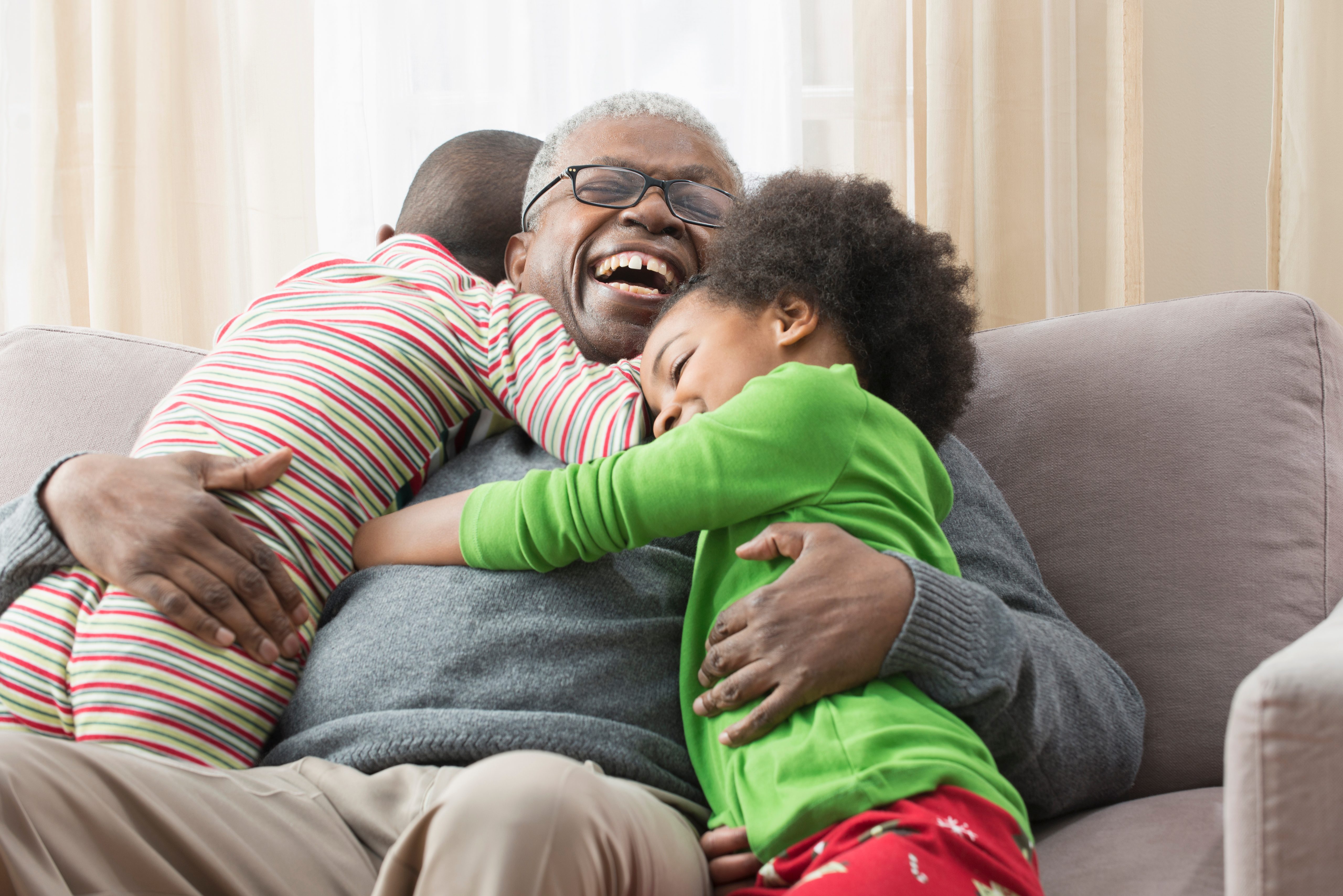 A person smiling while two children hug them tightly on a comfortable couch in a warmly lit room.