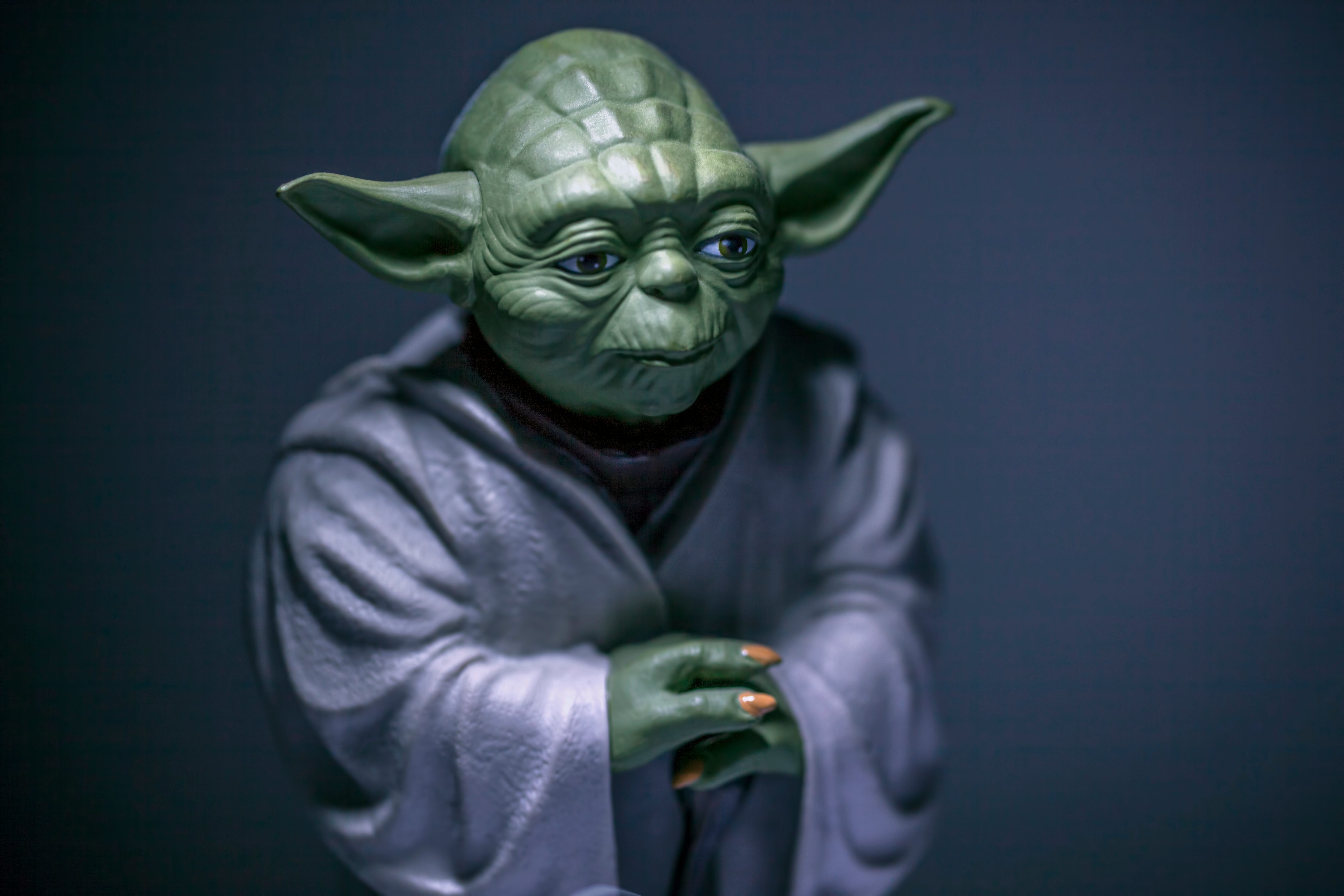 Green figure stands quietly, hands folded, wearing a textured robe against a dark, solid background.