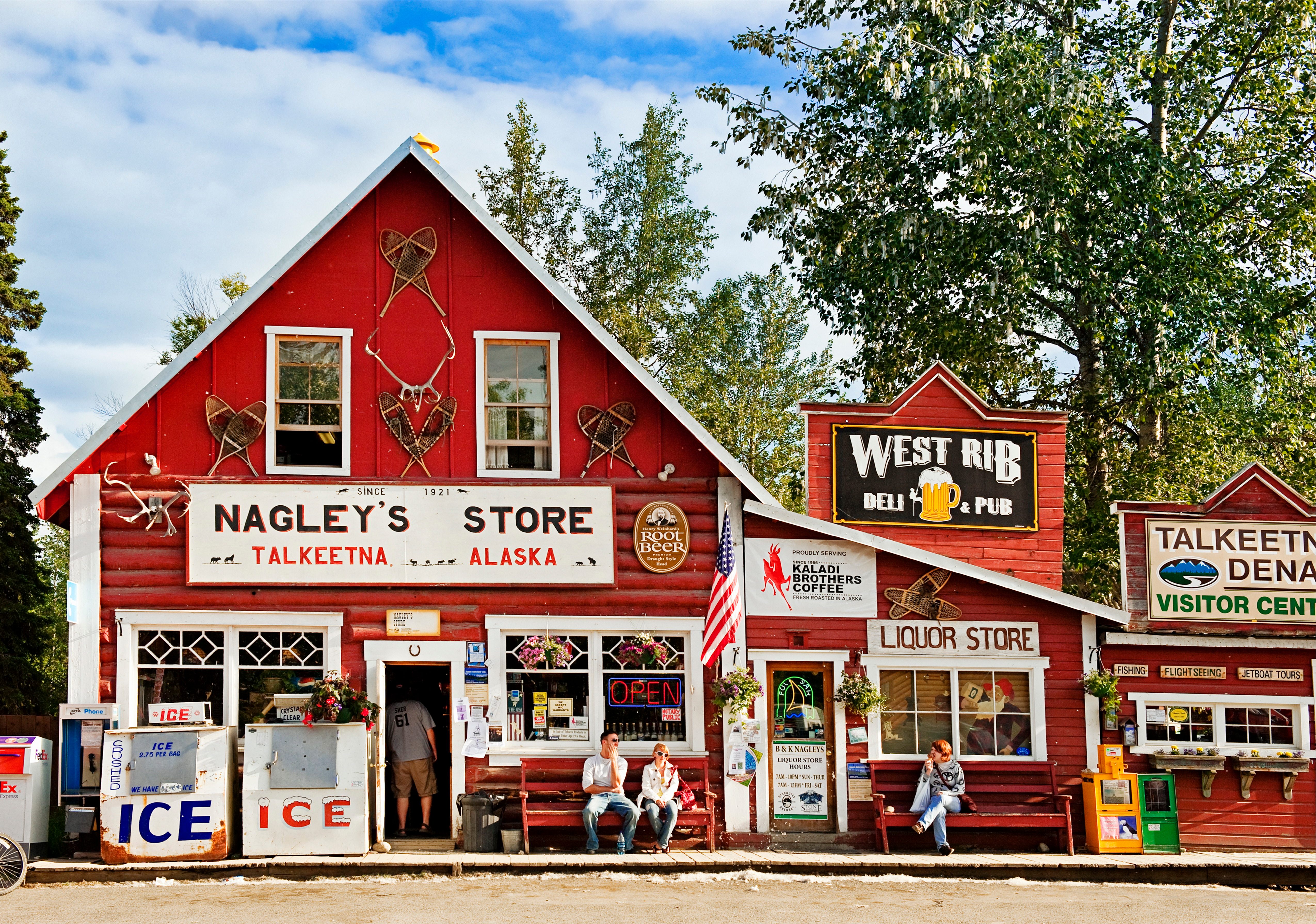 A red, rustic store displays antlers and signs, with people sitting outside. It's surrounded by trees under a cloudy sky. Text reads: "Nagley's Store."
