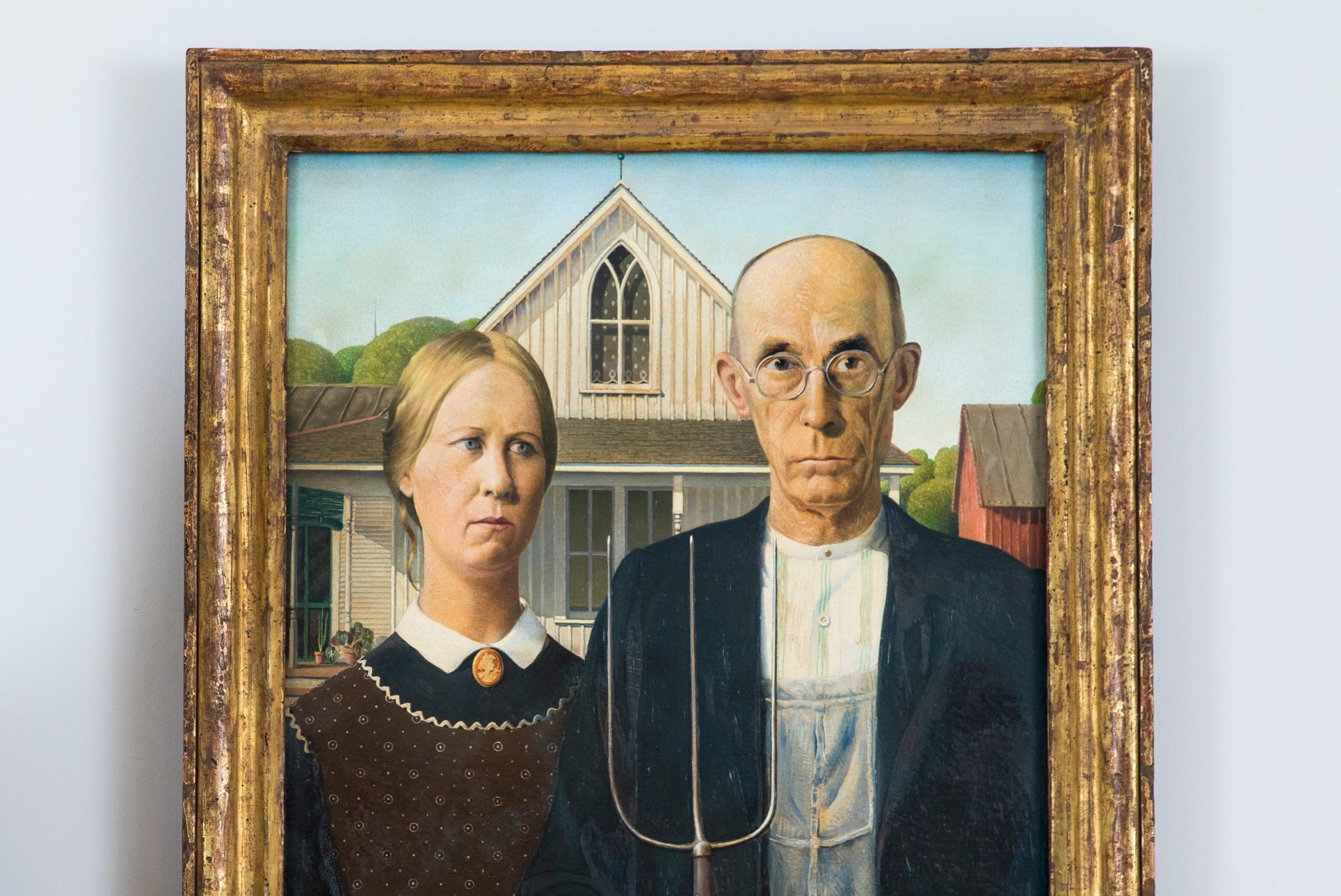 A stern man and woman stand in front of a farmhouse, the man holding a pitchfork, with a Gothic window in the background.