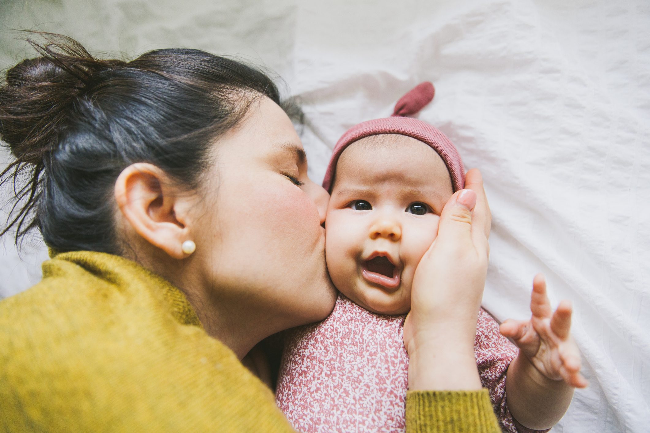 Woman kisses baby's cheek, holding its face gently; baby wears a pink hat, lying on a white blanket.