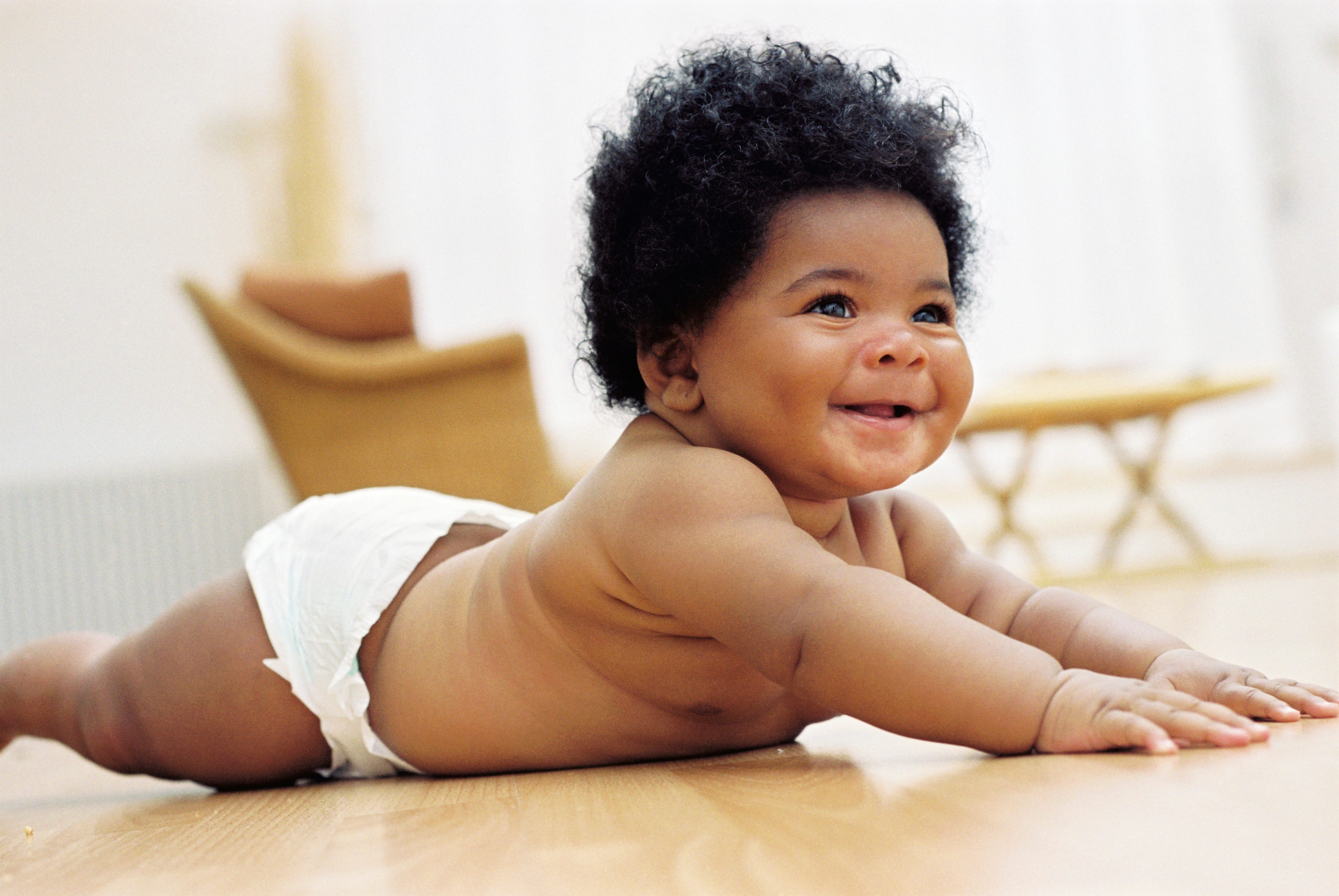 Baby smiling, lying on a floor, wearing a diaper. Bright, softly focused room with wooden furniture blurred in the background.