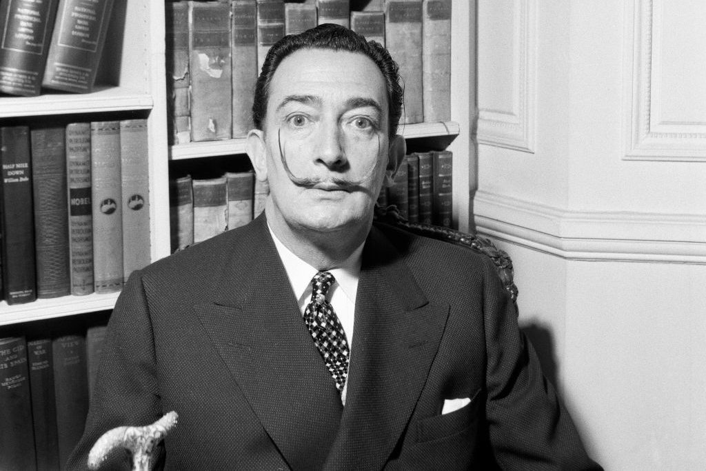 A man with a distinctive mustache sits in a library, surrounded by shelves of books, wearing a suit and tie, holding a cane.