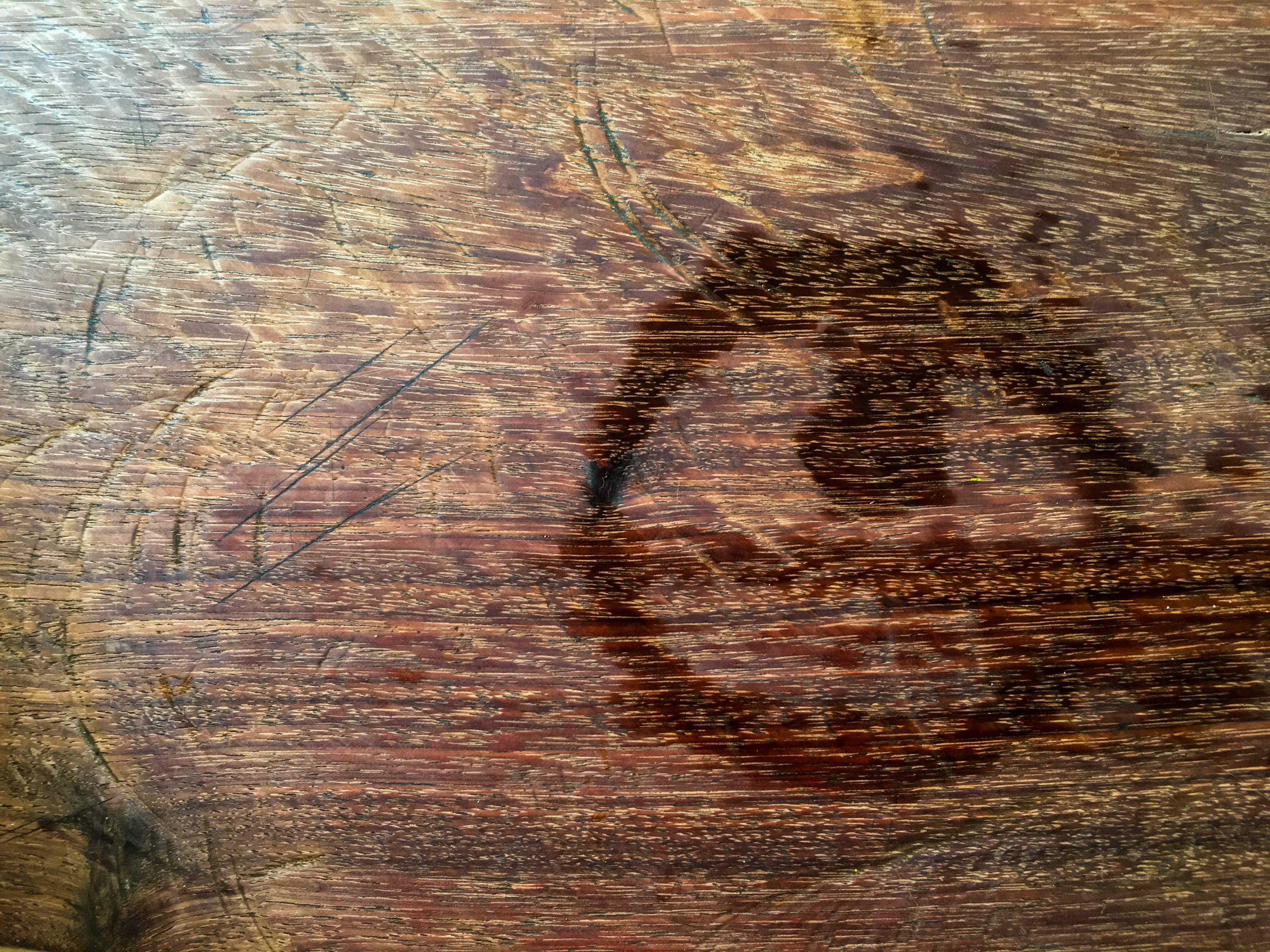 Circular stain forms on a scratched wooden surface, creating a dark ring amidst a backdrop of visible wood grain patterns.