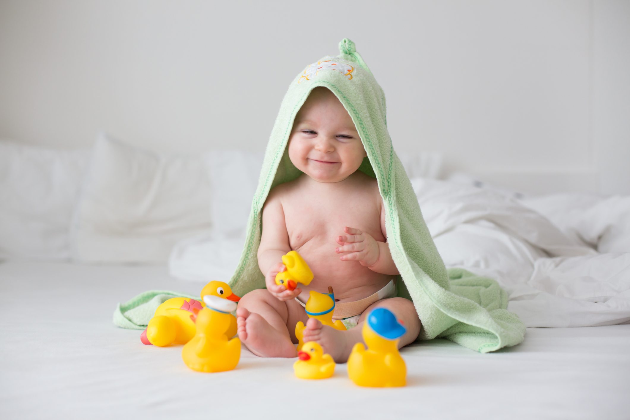 Baby smiles, sitting with yellow rubber ducks, wrapped in a green towel, on a white bed.