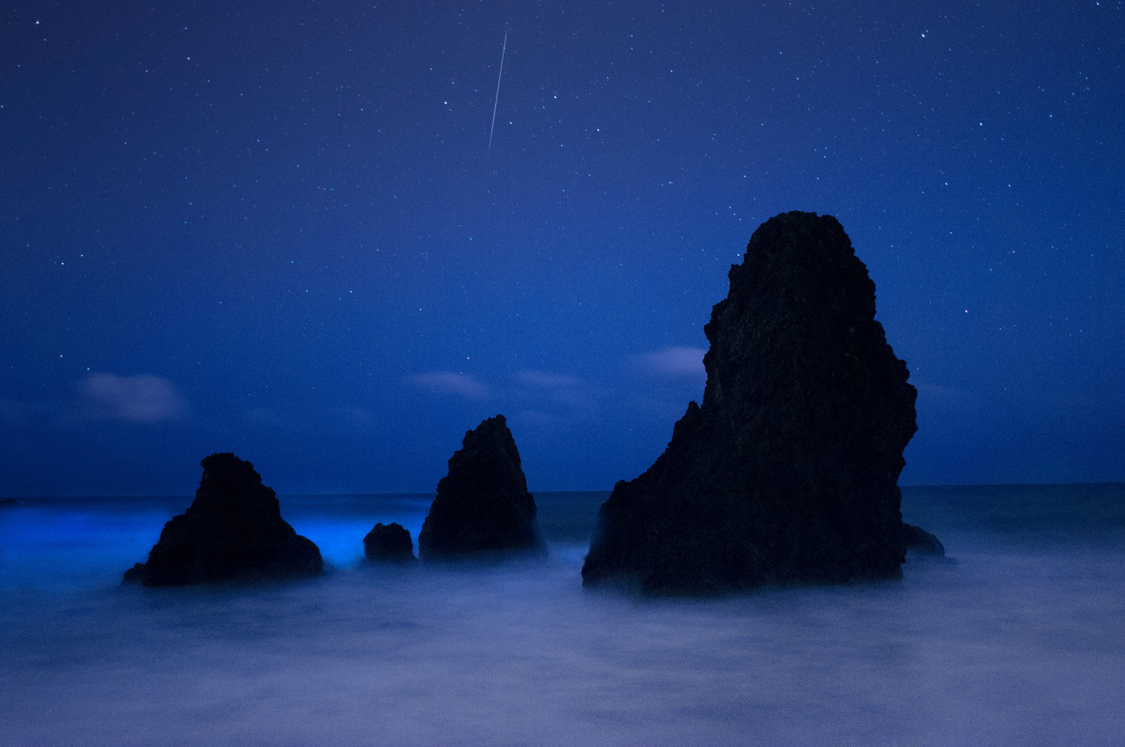 Rock formations rise from misty water under a starry sky, with a shooting star visible above in a nighttime coastal scene.