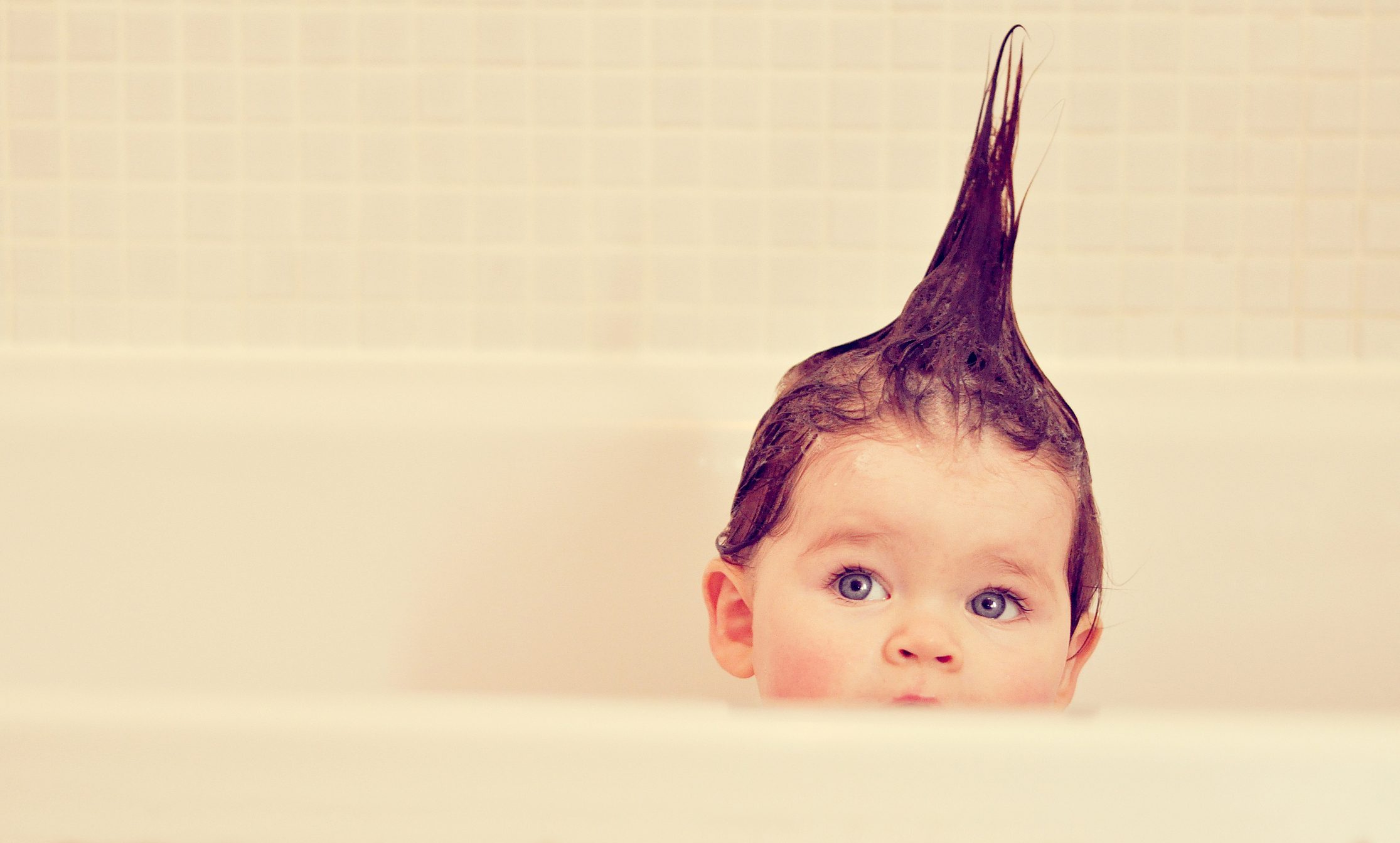 A baby peeks over the bathtub edge, hair styled into a playful point, surrounded by a tiled bathroom wall.