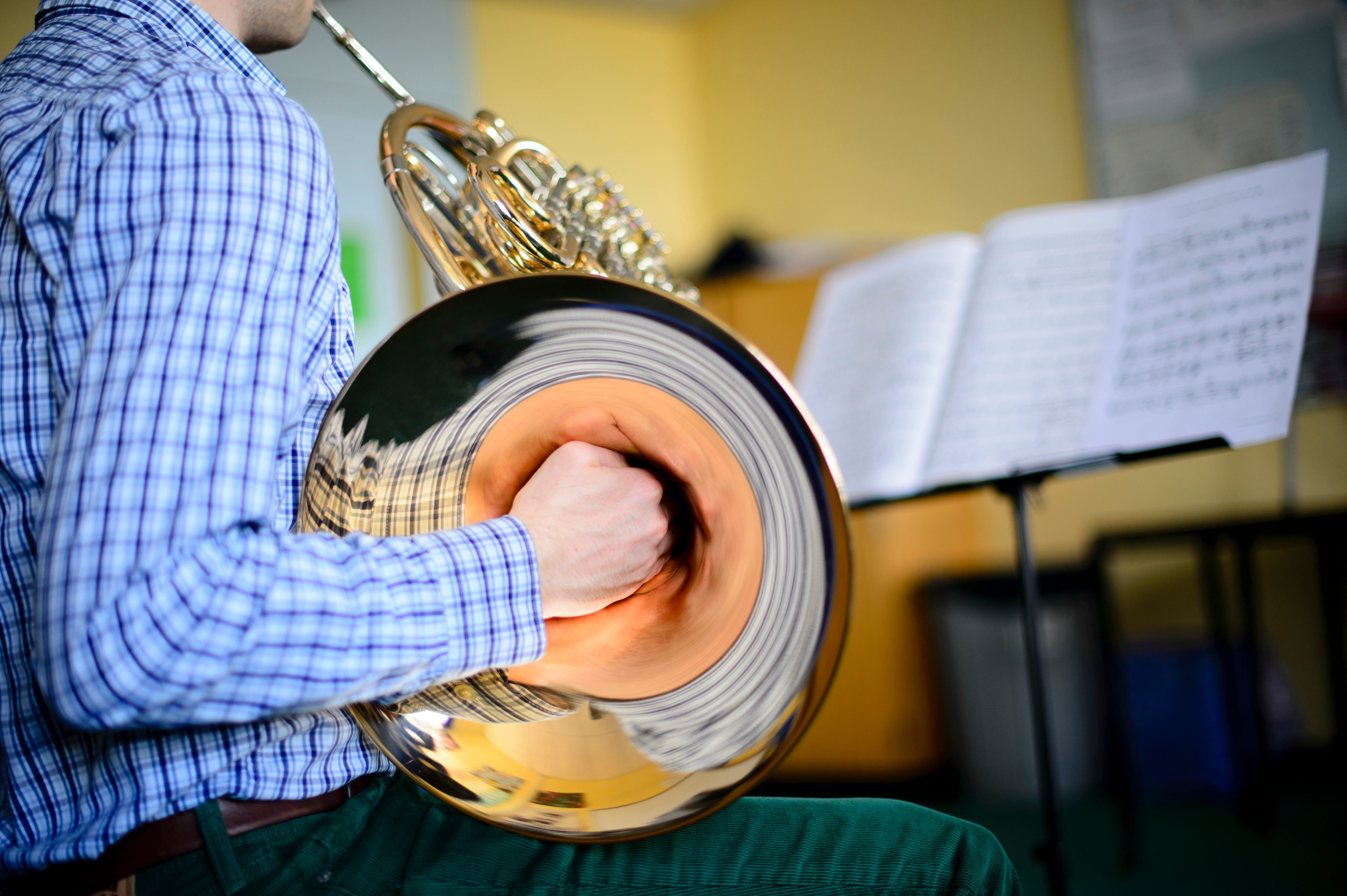 A person plays a French horn, wearing a checkered shirt, with sheet music on a stand in a brightly lit room.