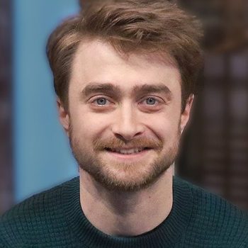 A person smiles in a close-up portrait, wearing a dark sweater, with a blurred indoor setting in the background.