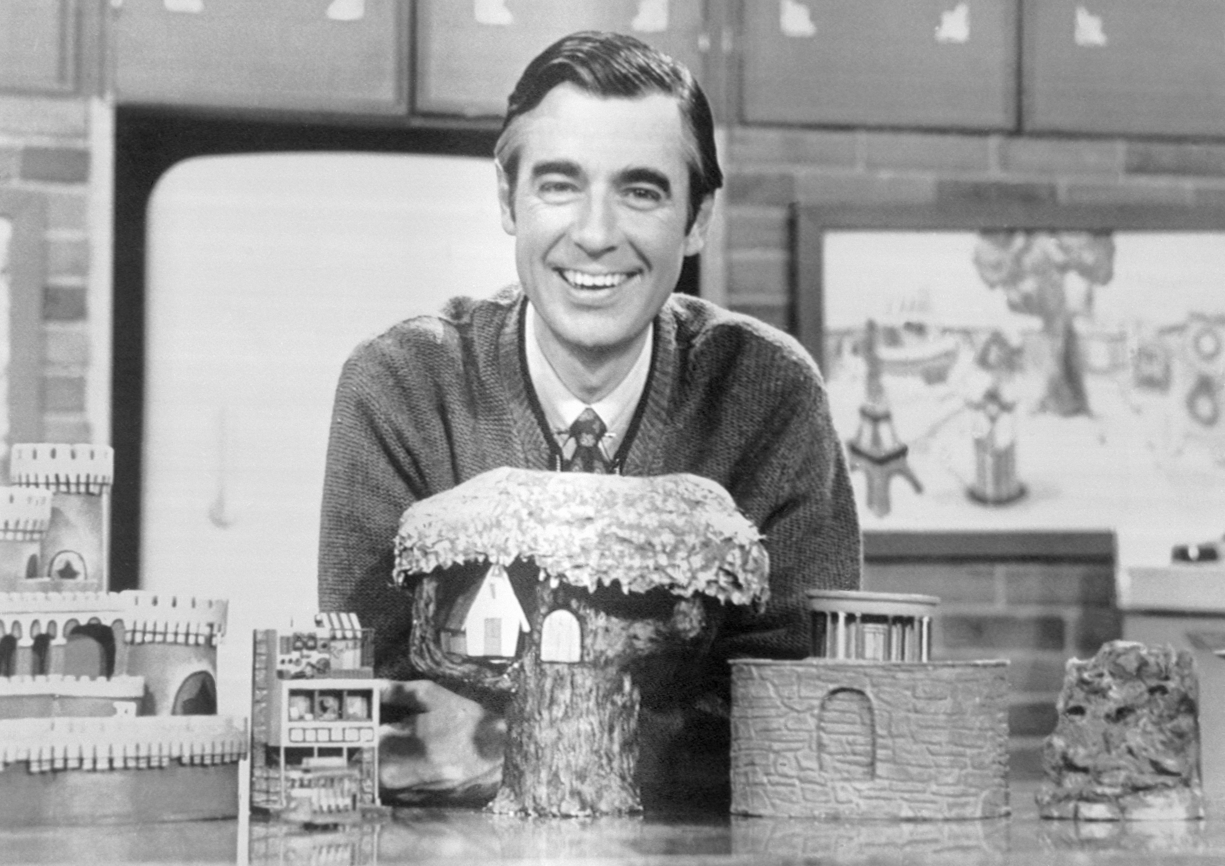 A person smiles warmly behind miniature model buildings on a desk, set in a cozy room with a brick wall and artwork in the background.