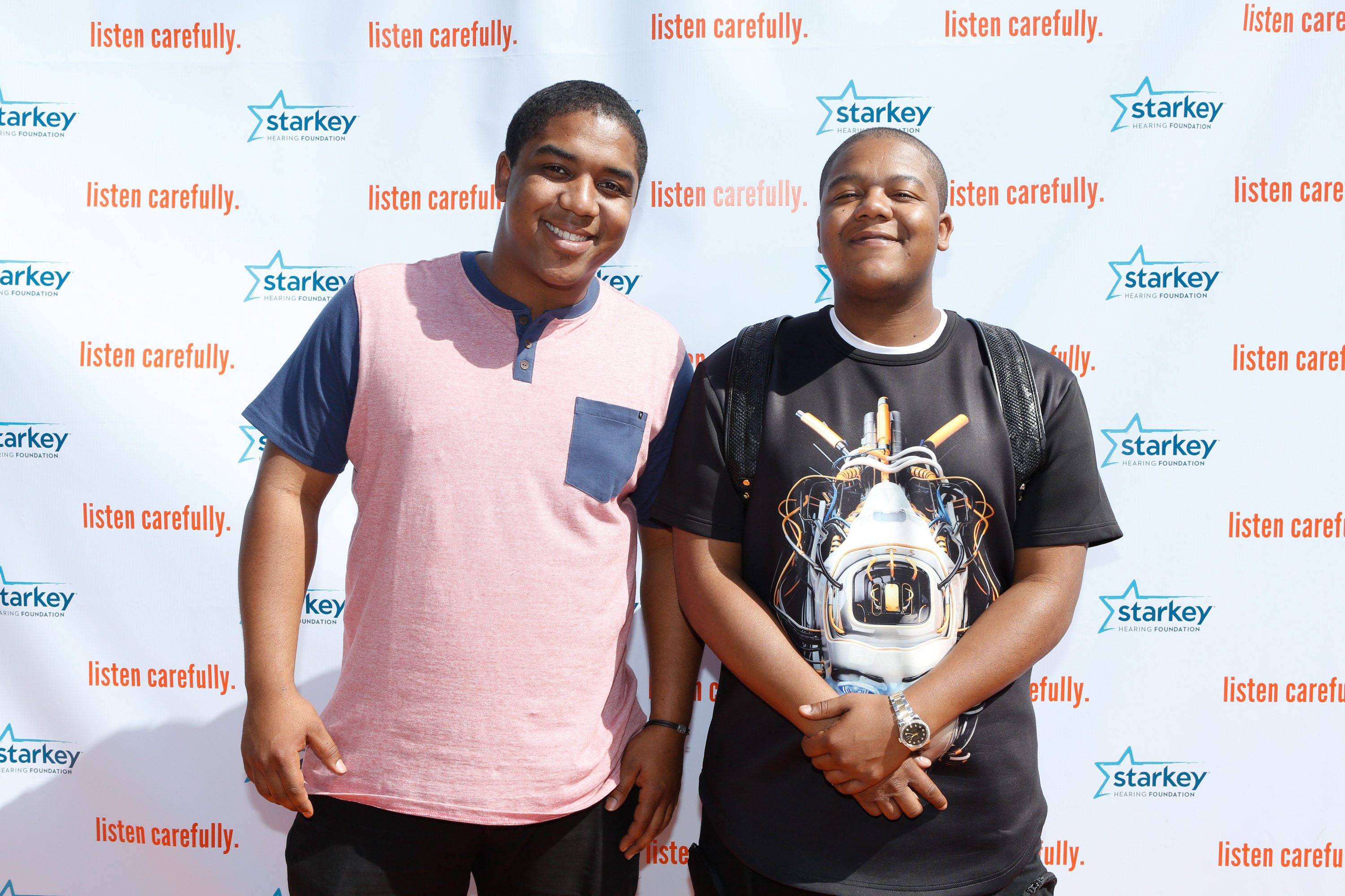 Two people smiling, standing together against a backdrop with "listen carefully" and "starkey hearing foundation" logos repeatedly printed on it.