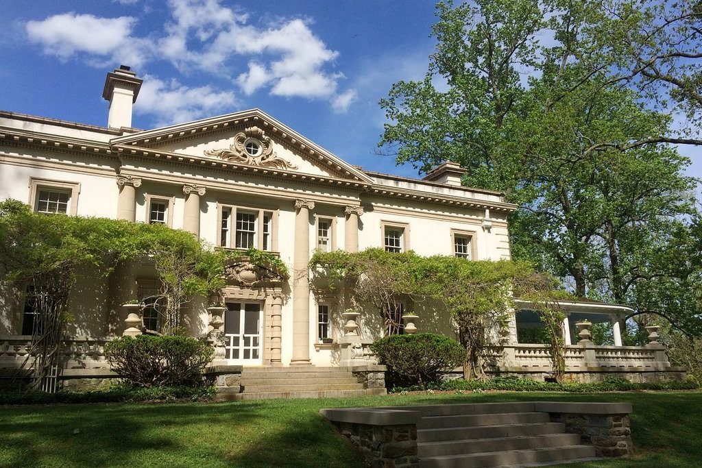 A stately mansion sits beneath a blue sky, surrounded by lush greenery and climbing vines, creating a serene and elegant landscape.