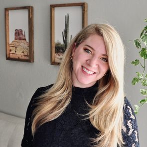 A person smiling, standing indoors; behind them are two framed desert-themed photographs and a plant with green leaves to the right.