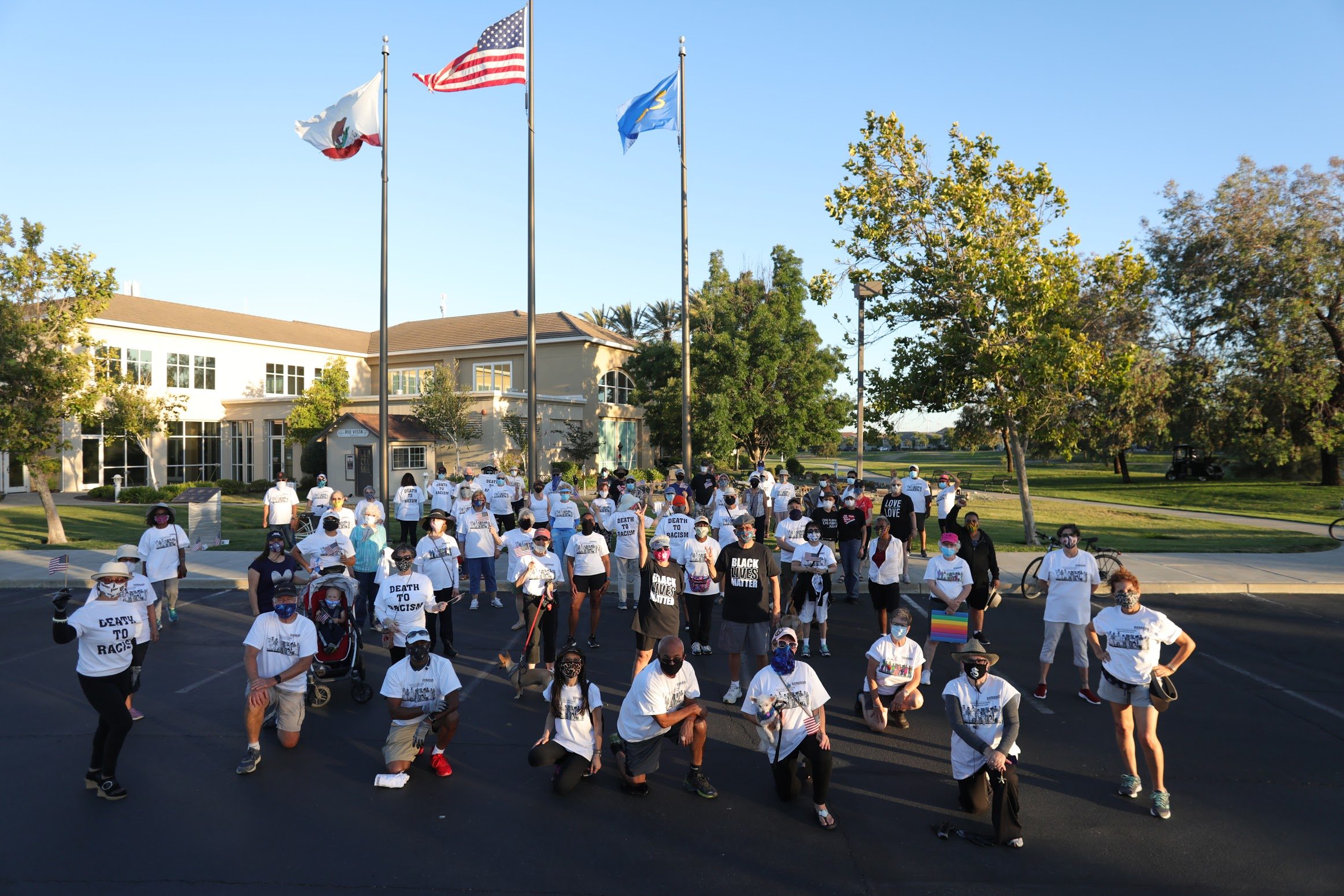 Crowd gathers peacefully, wearing protest shirts, in a park near flagpoles with three flags, during daylight.