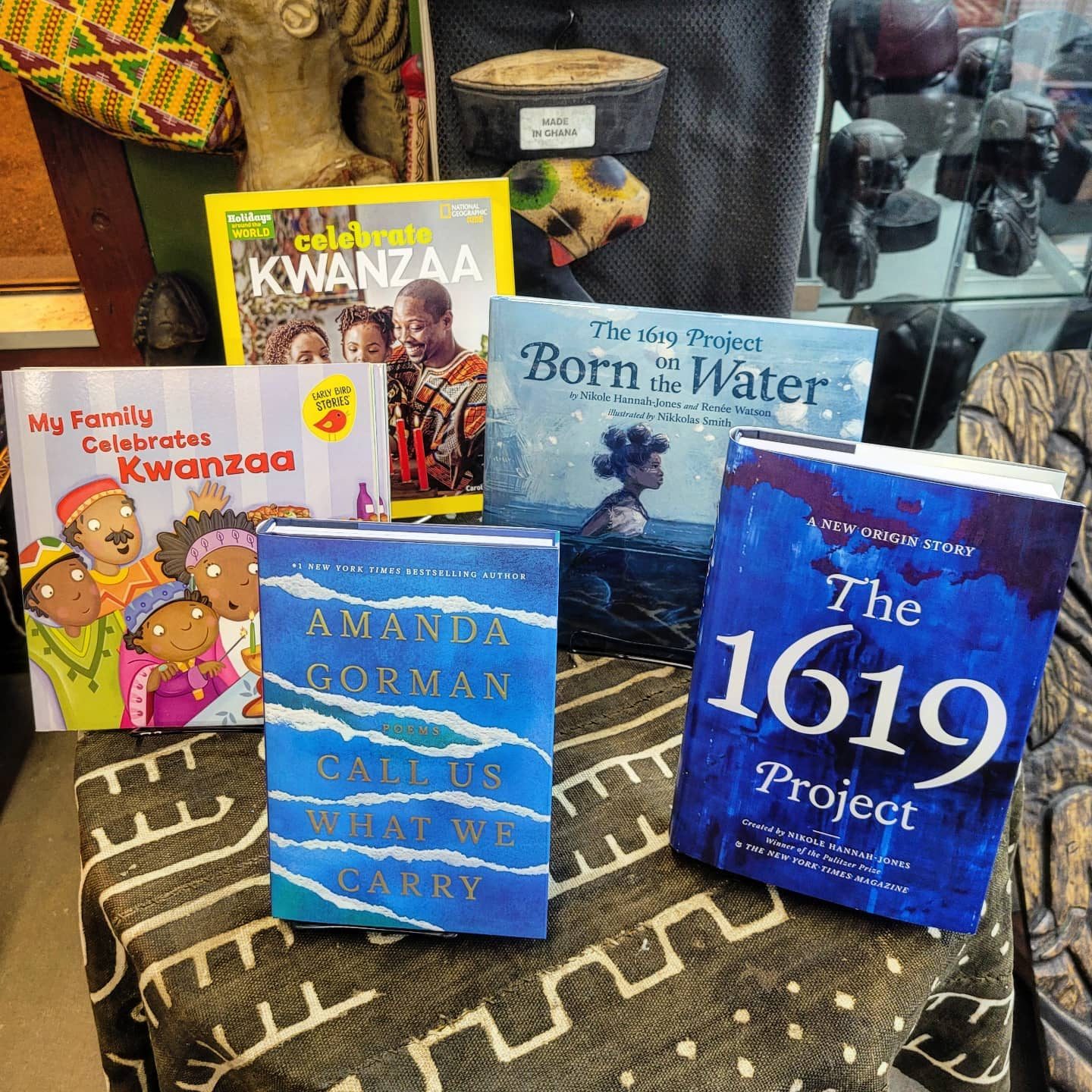 Books are displayed upright on a patterned tablecloth, featuring titles on Kwanzaa, history, and poetry. Background includes an African art piece and textiles.