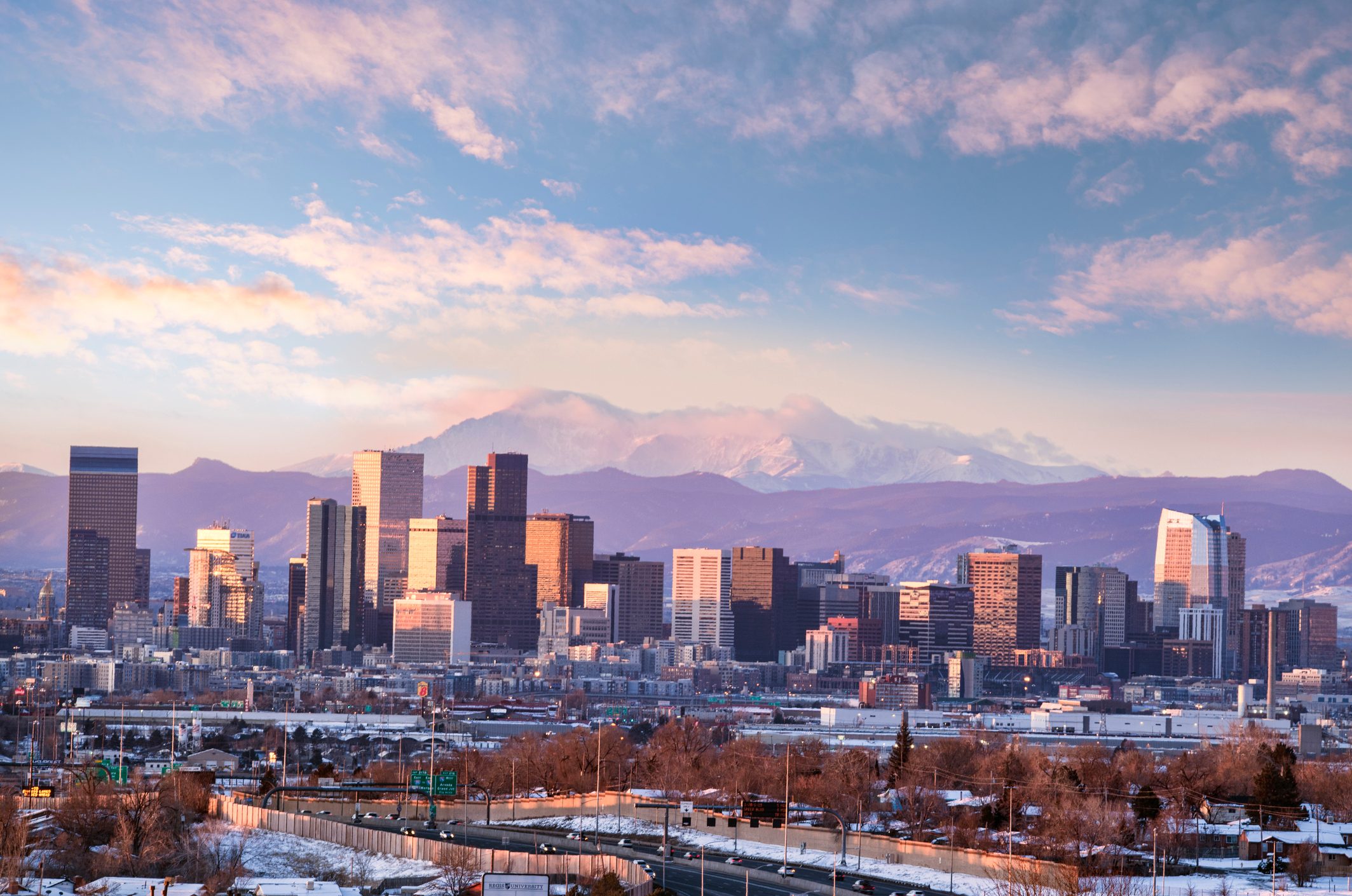 Skyscrapers bathe in sunset light, set against a mountainous backdrop with scattering clouds. The foreground features roads and sparse winter vegetation.