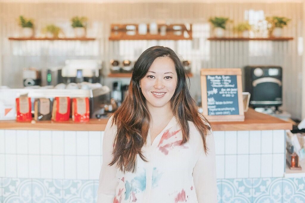 A woman stands smiling at a coffee shop counter, surrounded by coffee bags and a chalkboard menu, with plants on shelves behind her.