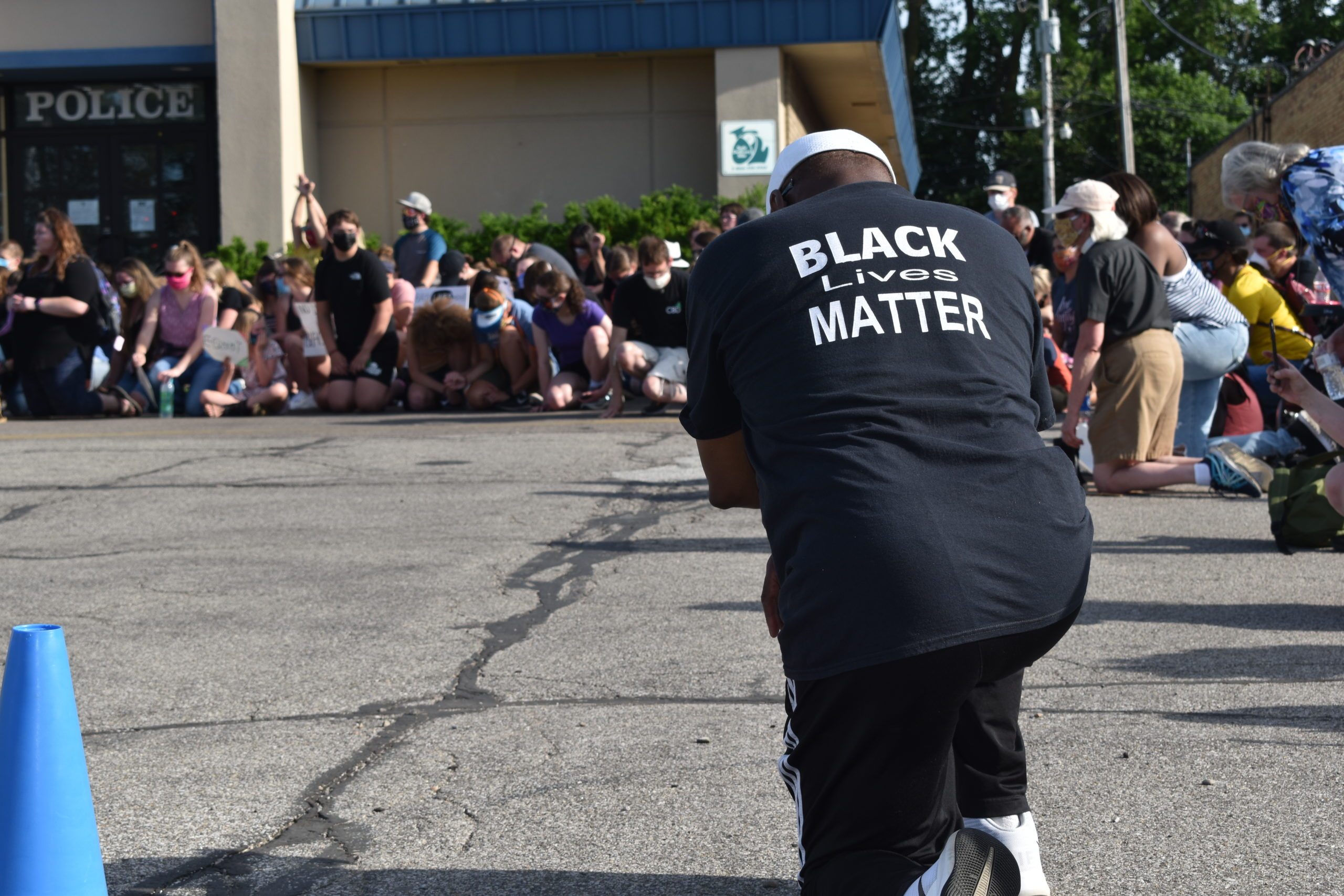 Person kneels wearing "Black Lives Matter" shirt, facing a crowd kneeling near a building with visible "POLICE" sign.