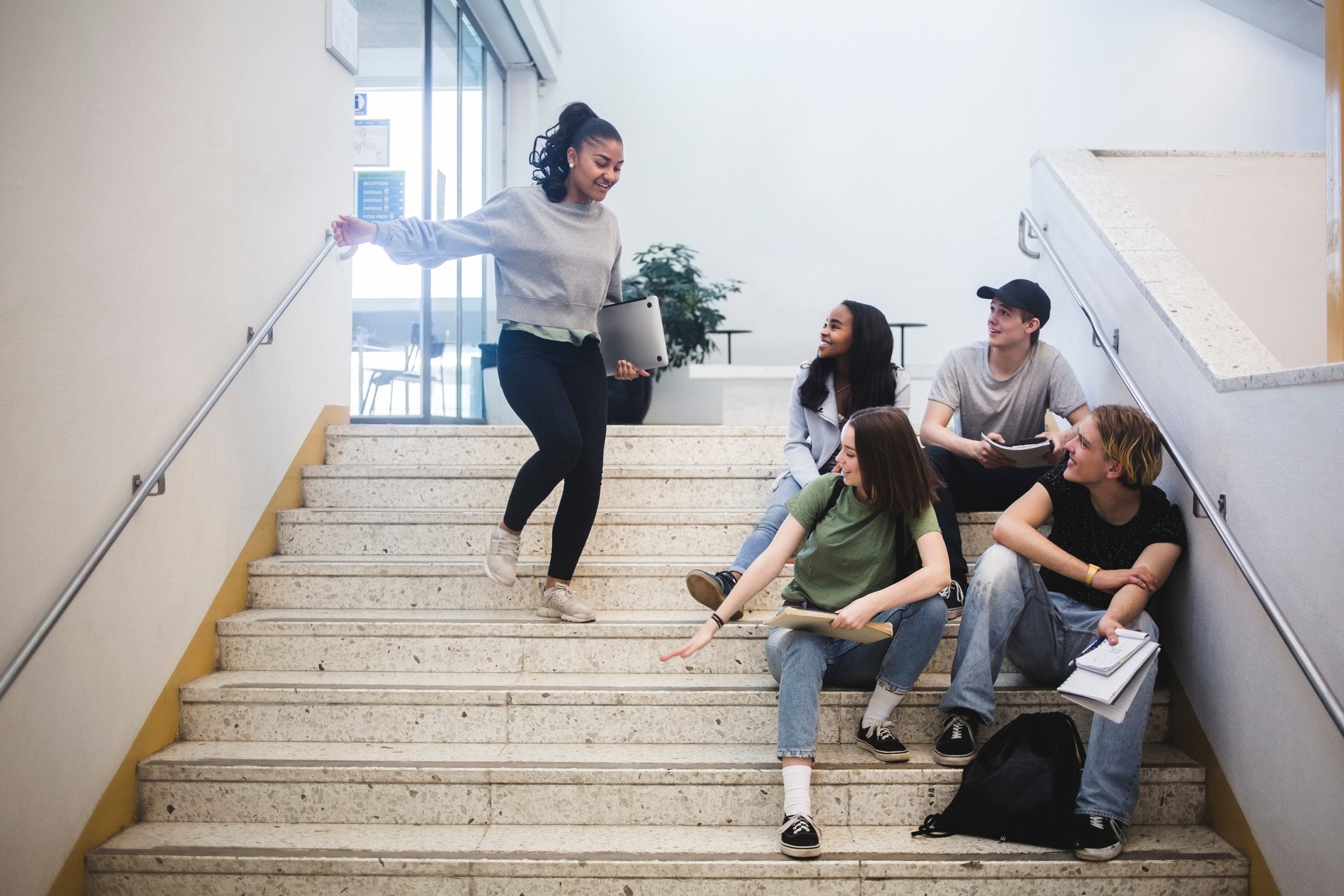 Students sit on stairs, engaging with a walking peer. Indoors, bright setting, casual atmosphere.