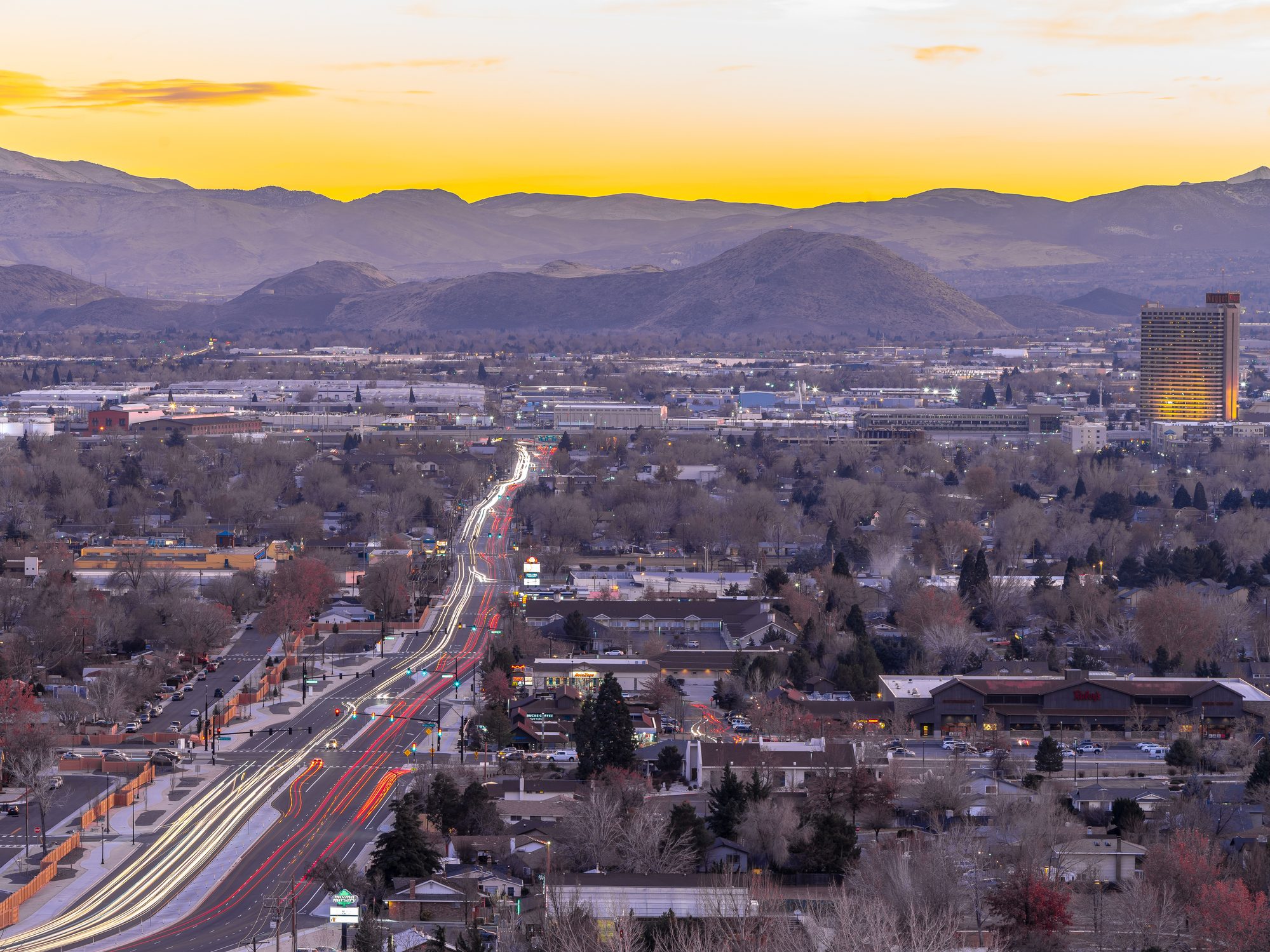Cityscape shows a road with light trails, surrounded by buildings and trees, against a backdrop of mountains and a yellow sky at dusk.