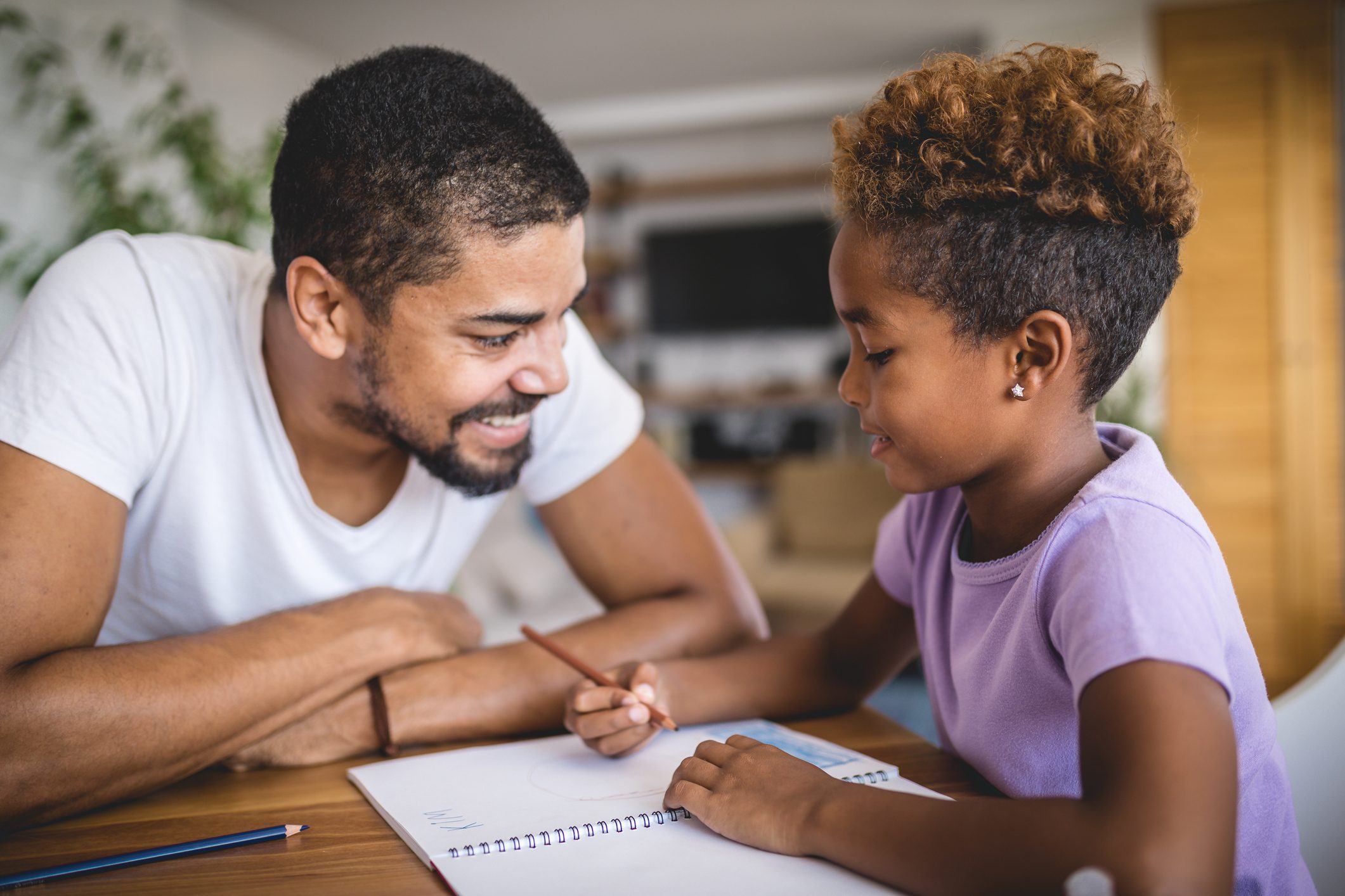 Child draws with pencil while adult watches with a smile, seated at a wooden table in a cozy living room setting.