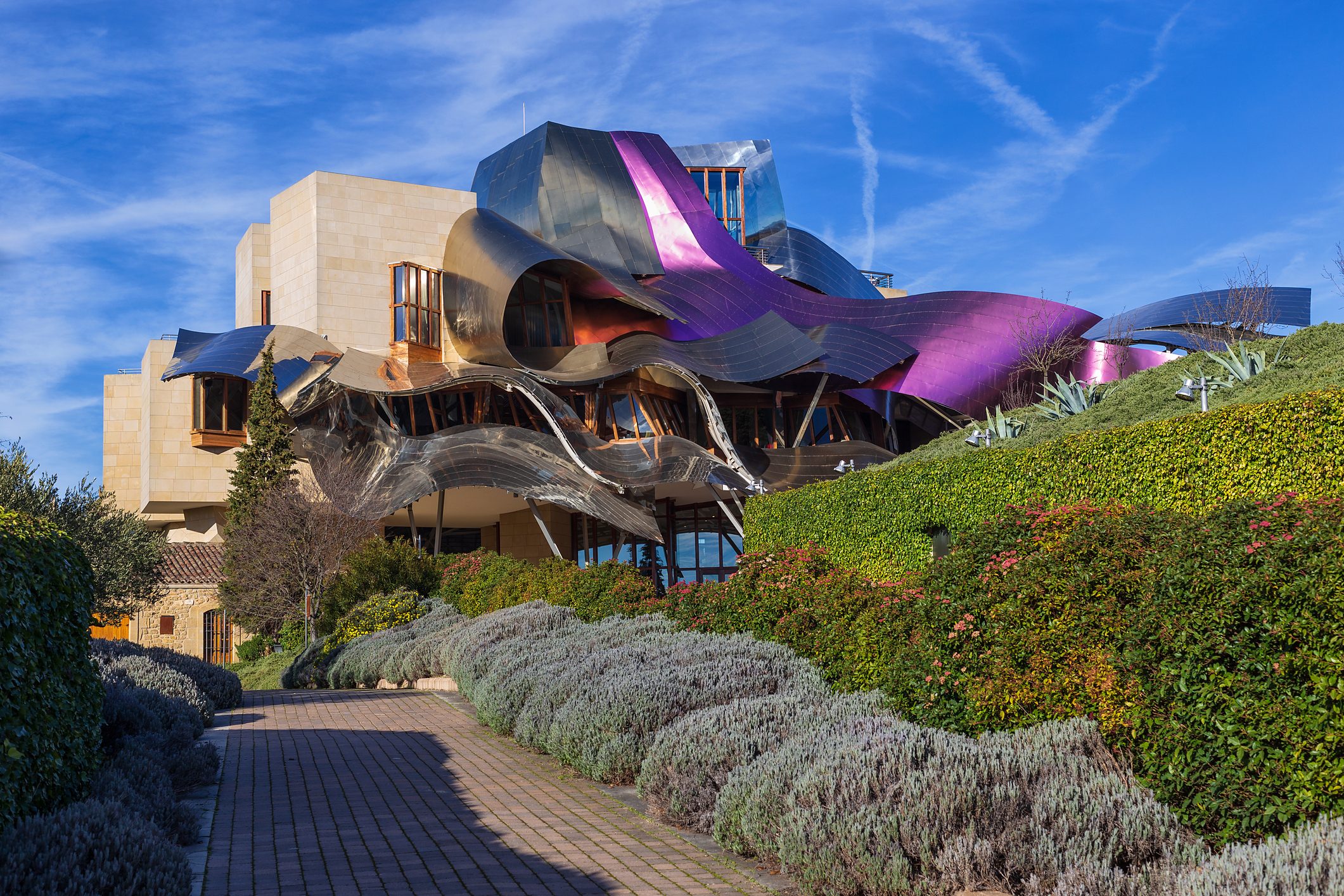 Building with metallic, wavy panels reflects sunlight, surrounded by lush green shrubs and colorful plants under a clear blue sky.