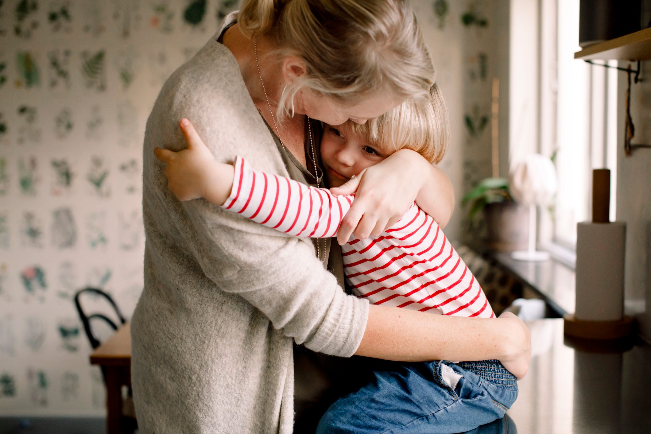 A child in a red-striped shirt hugs an adult in a cozy kitchen with plant-patterned wallpaper.