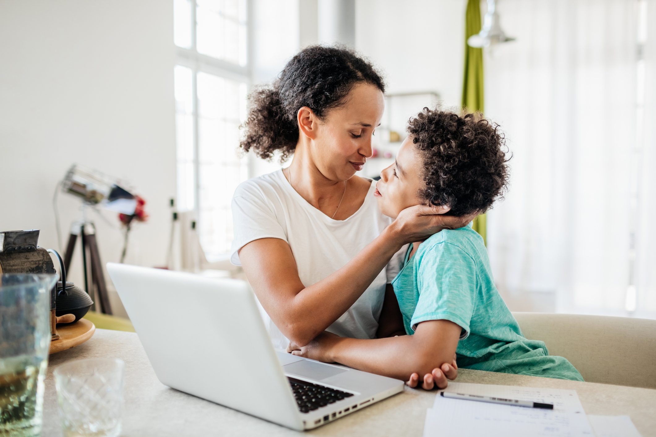 Woman embracing child gently, seated at table with laptop, bright room with large windows and modern decor in the background.