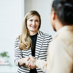 A woman in a striped blazer smiles while shaking hands with another person in an office setting.